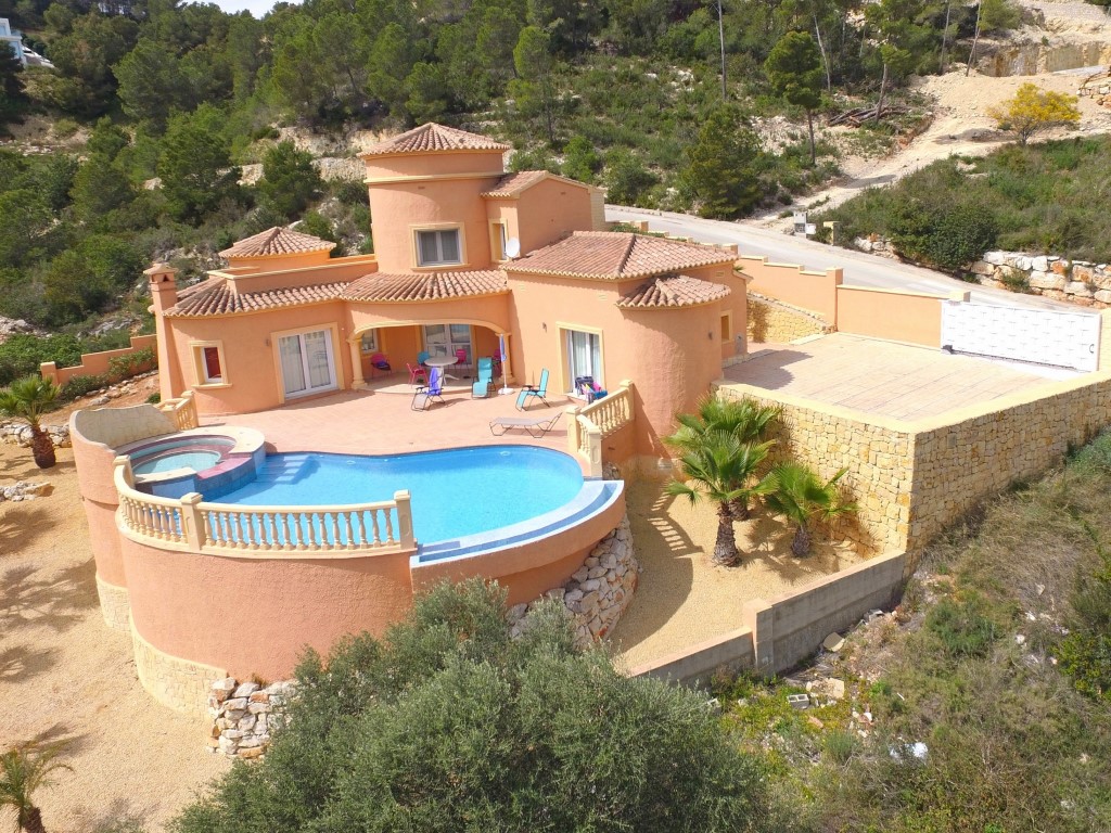 Elevated view of the villa featuring a curved infinity pool with integrated jacuzzi, terracotta terrace, and a spacious paved parking area enclosed by stone walls.