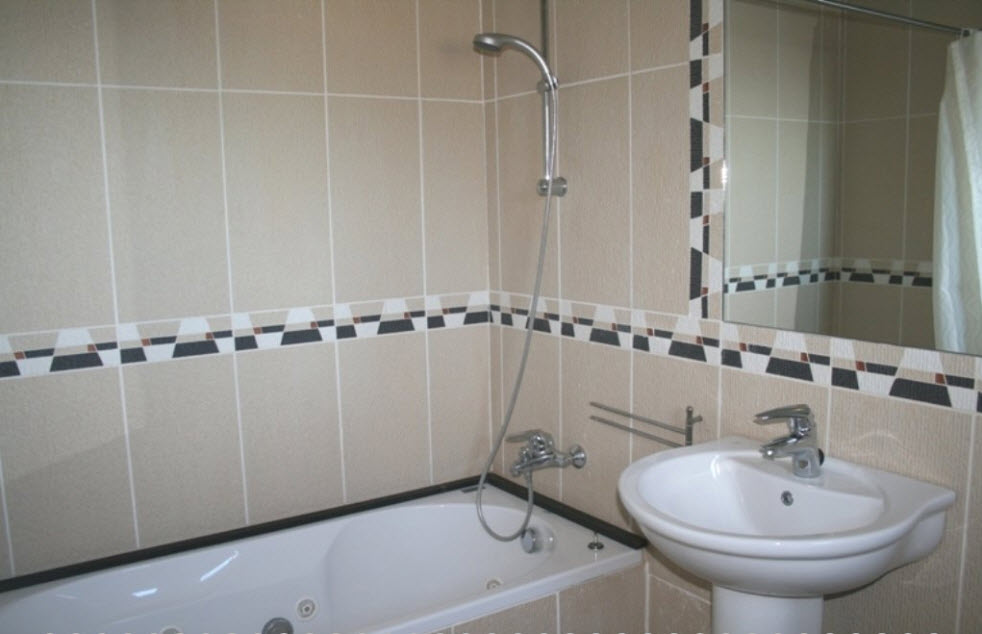 En-suite bathroom featuring a whirlpool bathtub, pedestal sink, and beige wall tiles accented by a geometric mosaic border and chrome fixtures.