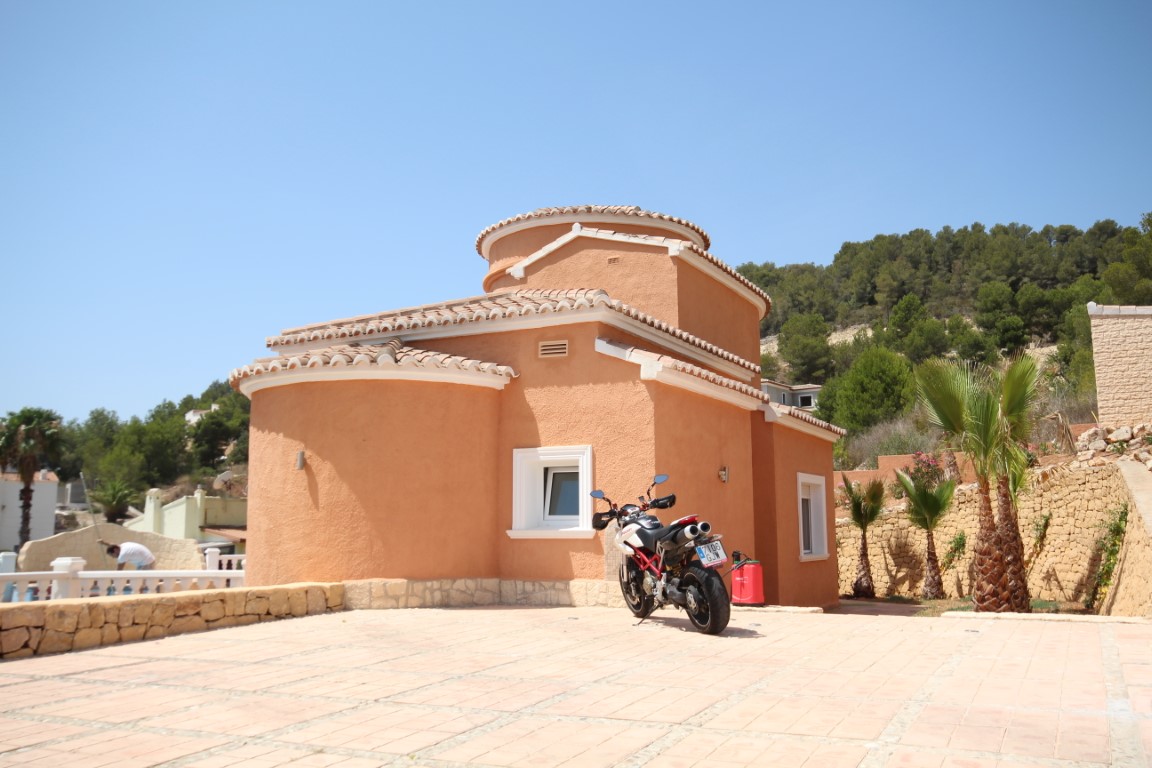 Exterior view of the villa featuring curved terracotta walls, a spacious paved parking area, and natural stone retaining walls set against a pine-covered hillside.