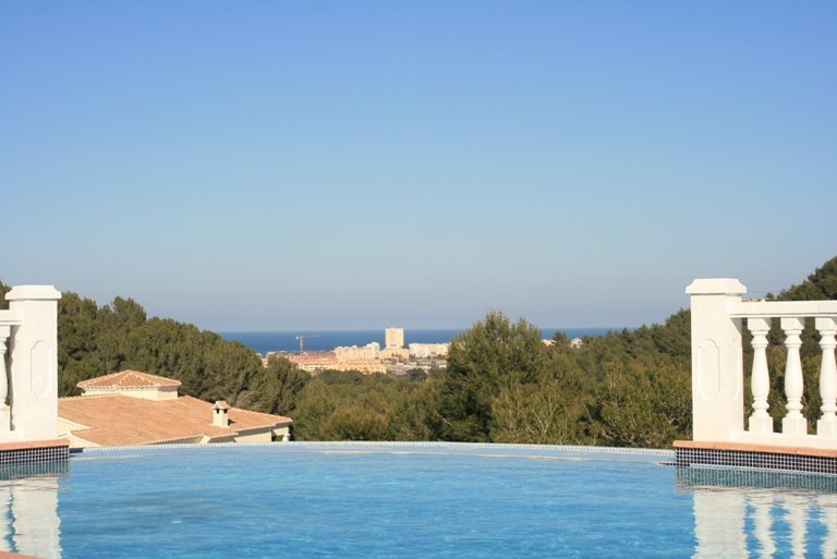 Infinity pool featuring blue mosaic tiling and white balustrades, overlooking a lush pine forest with distant views of the Mediterranean Sea.