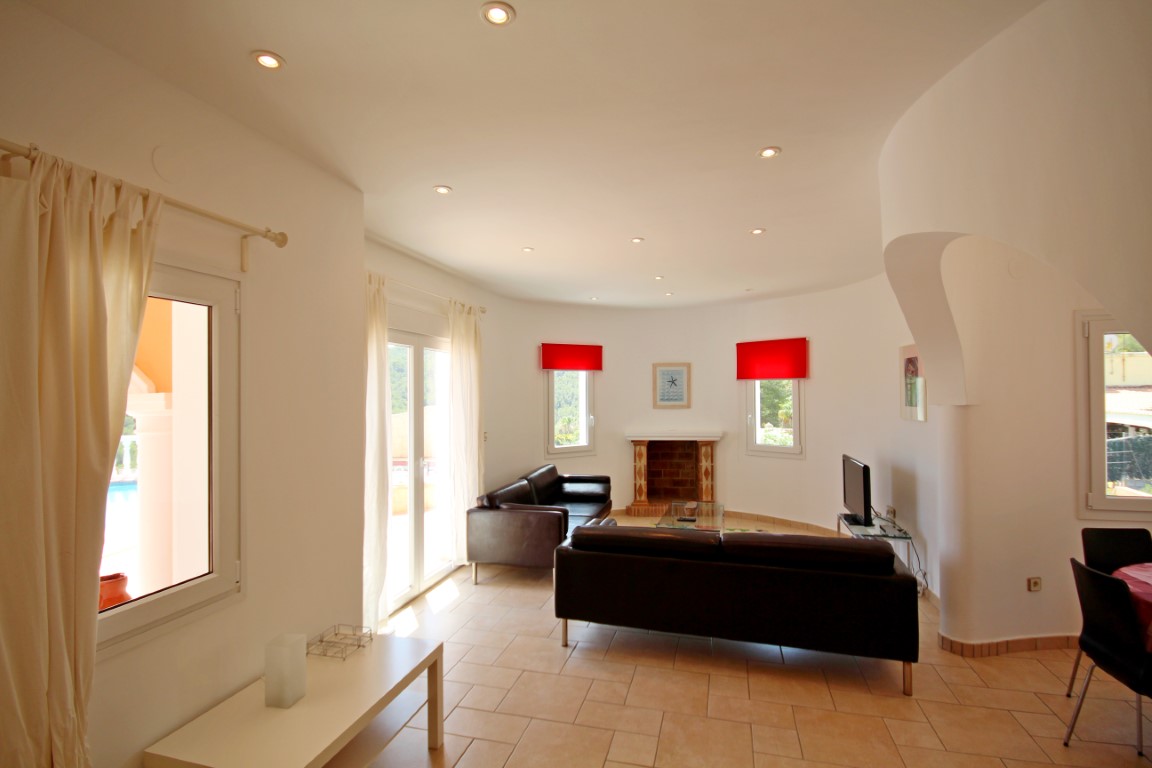 Living area featuring a brick fireplace, black leather sofas, and a curved ceiling with recessed spotlights. Large windows and doors offer direct access to the terrace.