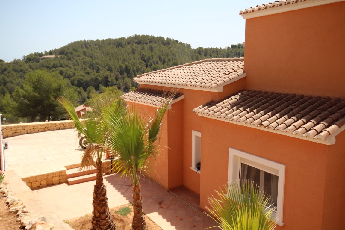 Mediterranean-style terracotta facade featuring traditional clay tile roofs, a paved patio with palm trees, and views of the surrounding green hillsides.