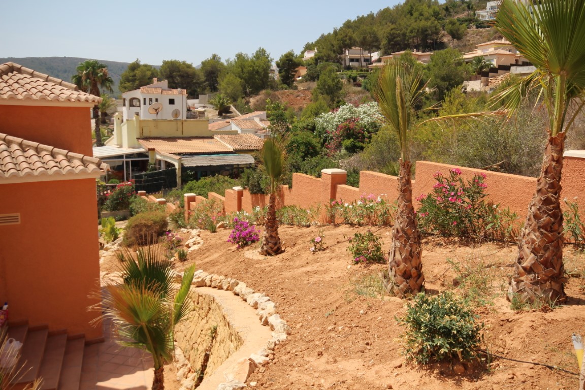 Terraced Mediterranean garden featuring young palm trees, flowering shrubs, and terracotta-colored perimeter walls overlooking the surrounding hillside.