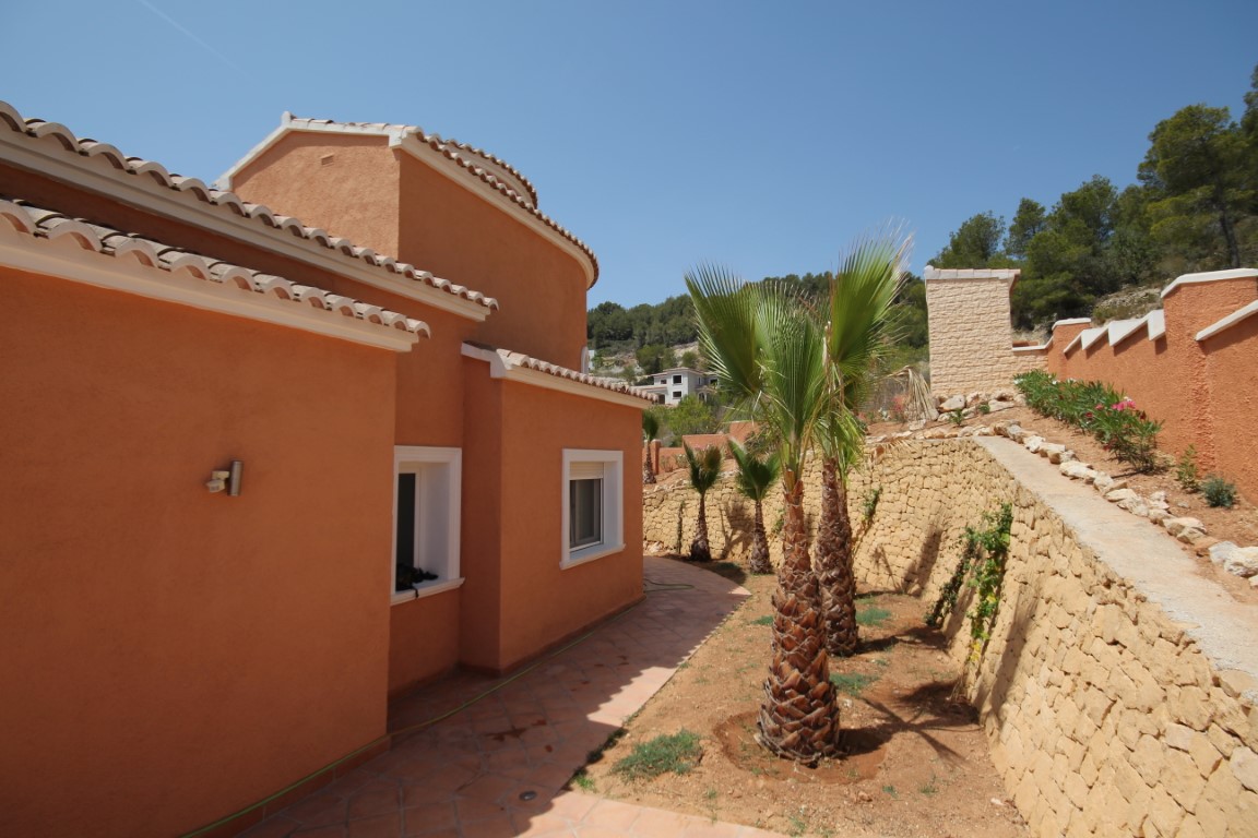 Terracotta-toned exterior featuring traditional clay tile roofing, landscaped with young palm trees and multi-level natural stone retaining walls.