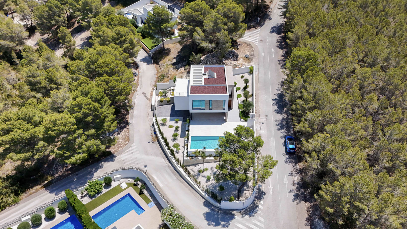 Aerial perspective of the modern white villa surrounded by a pine forest, featuring a turquoise pool, flat roof with solar panels, and minimalist landscaping.