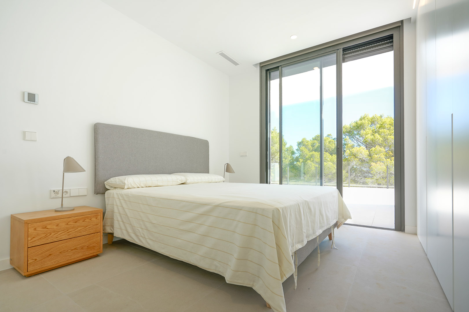 Bright bedroom featuring built-in wardrobes, large sliding glass doors, and direct access to a terrace overlooking the surrounding wooded area.