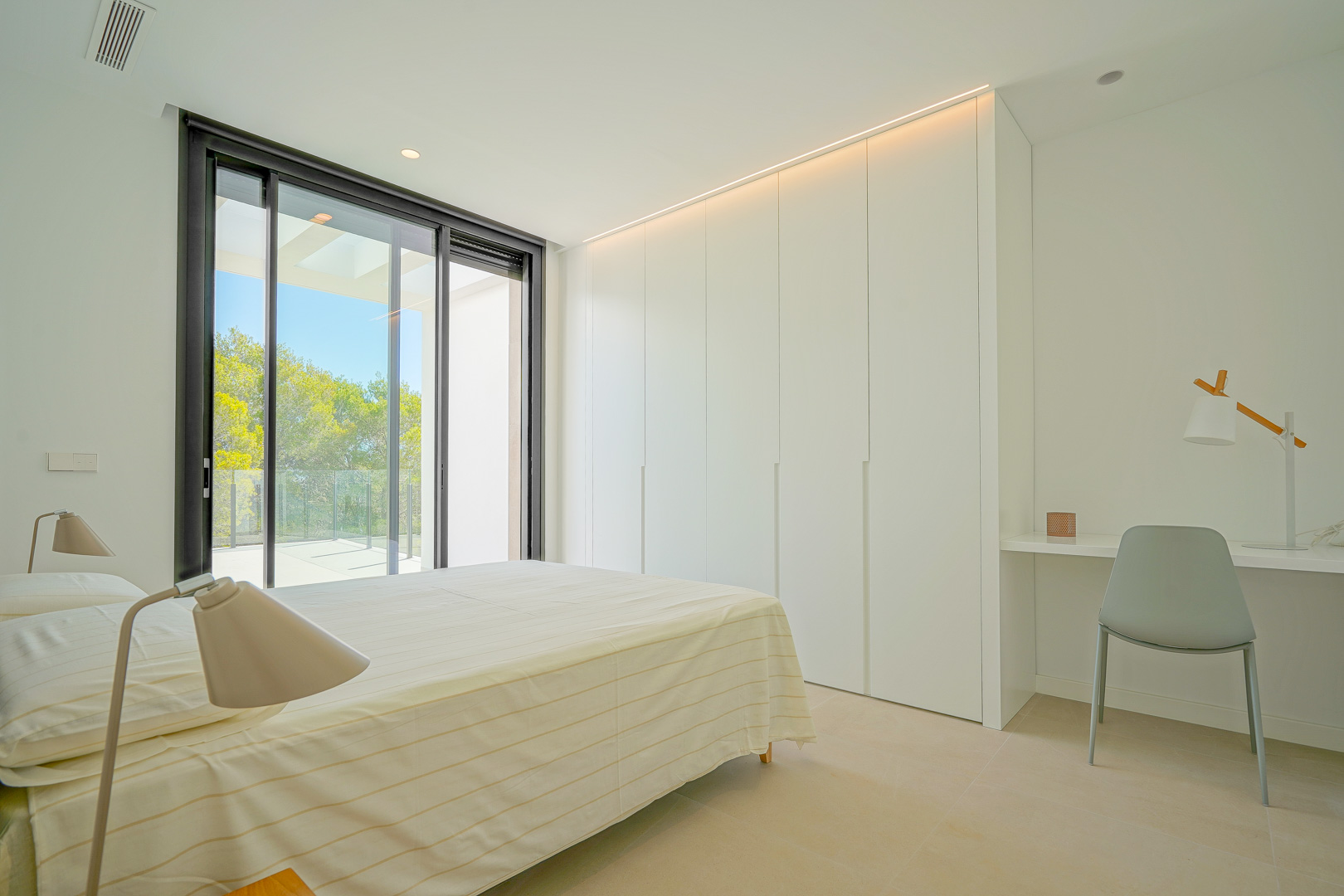Bright bedroom featuring floor-to-ceiling white wardrobes with LED strip lighting, a built-in workspace, and sliding doors opening to a private terrace.