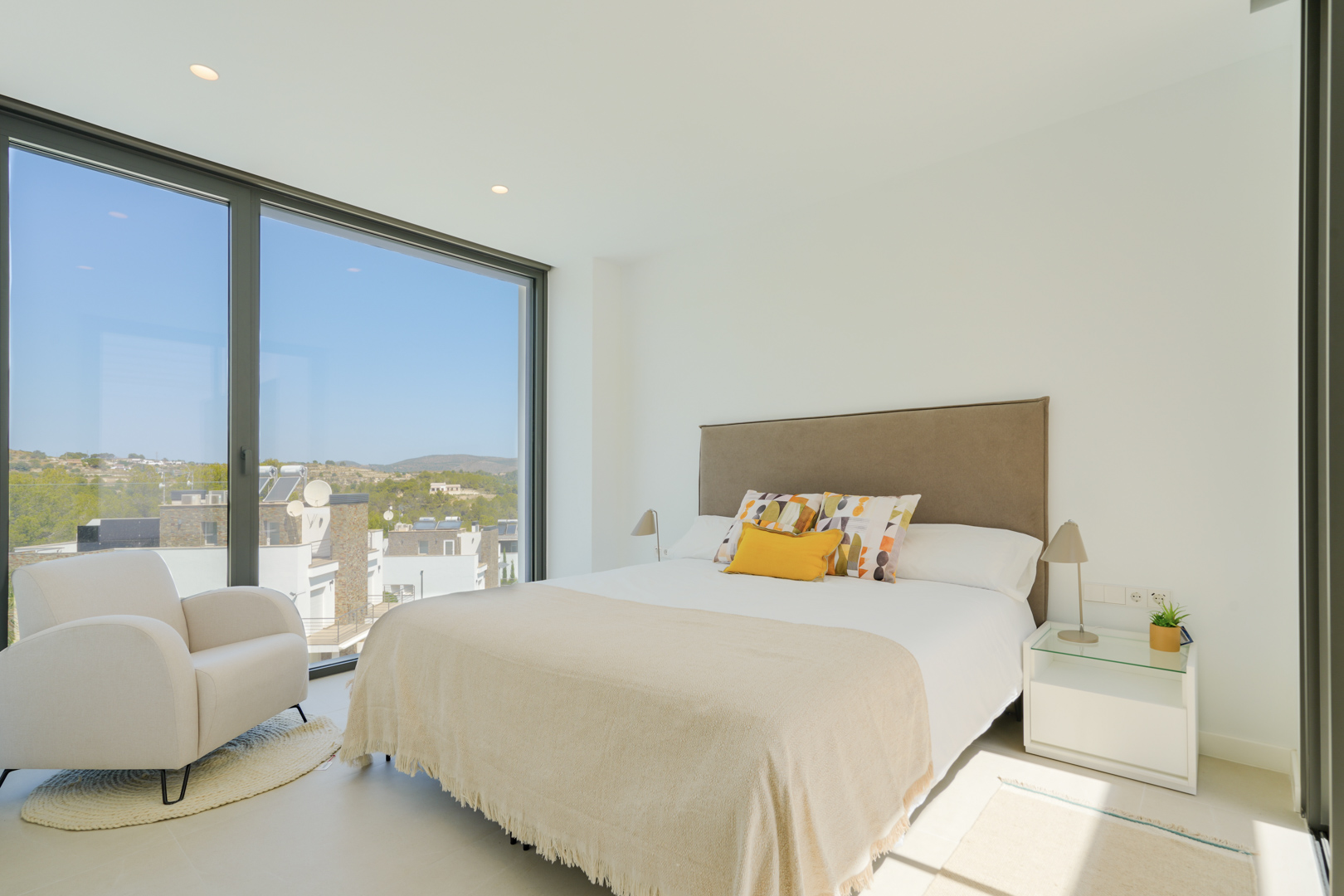 Bright bedroom featuring floor-to-ceiling windows, a double bed with a neutral upholstered headboard, and a minimalist armchair overlooking the valley views.