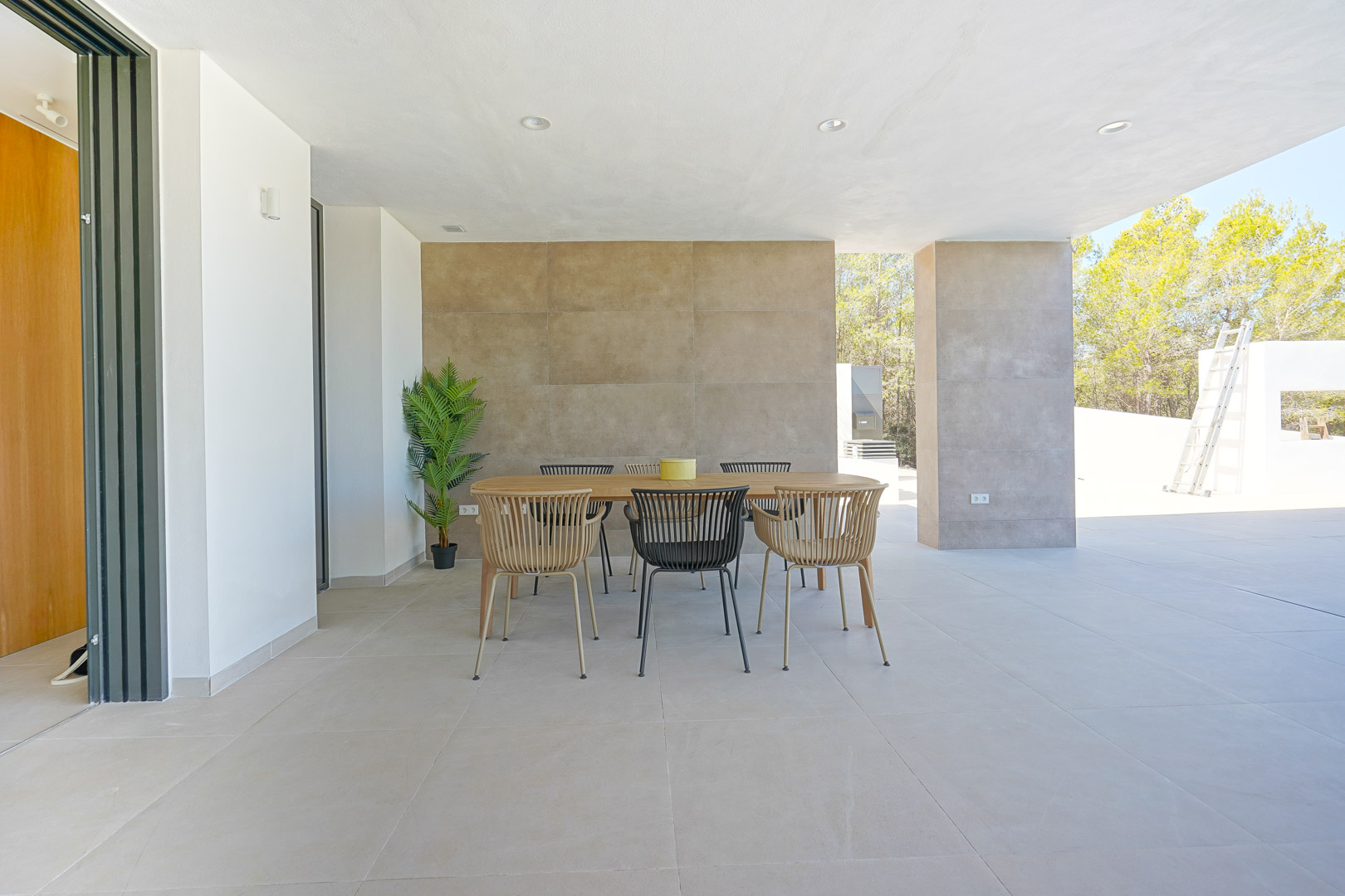 Covered terrace featuring an outdoor dining area with a wooden table, textured feature wall, and large-format floor tiles overlooking the surrounding trees.