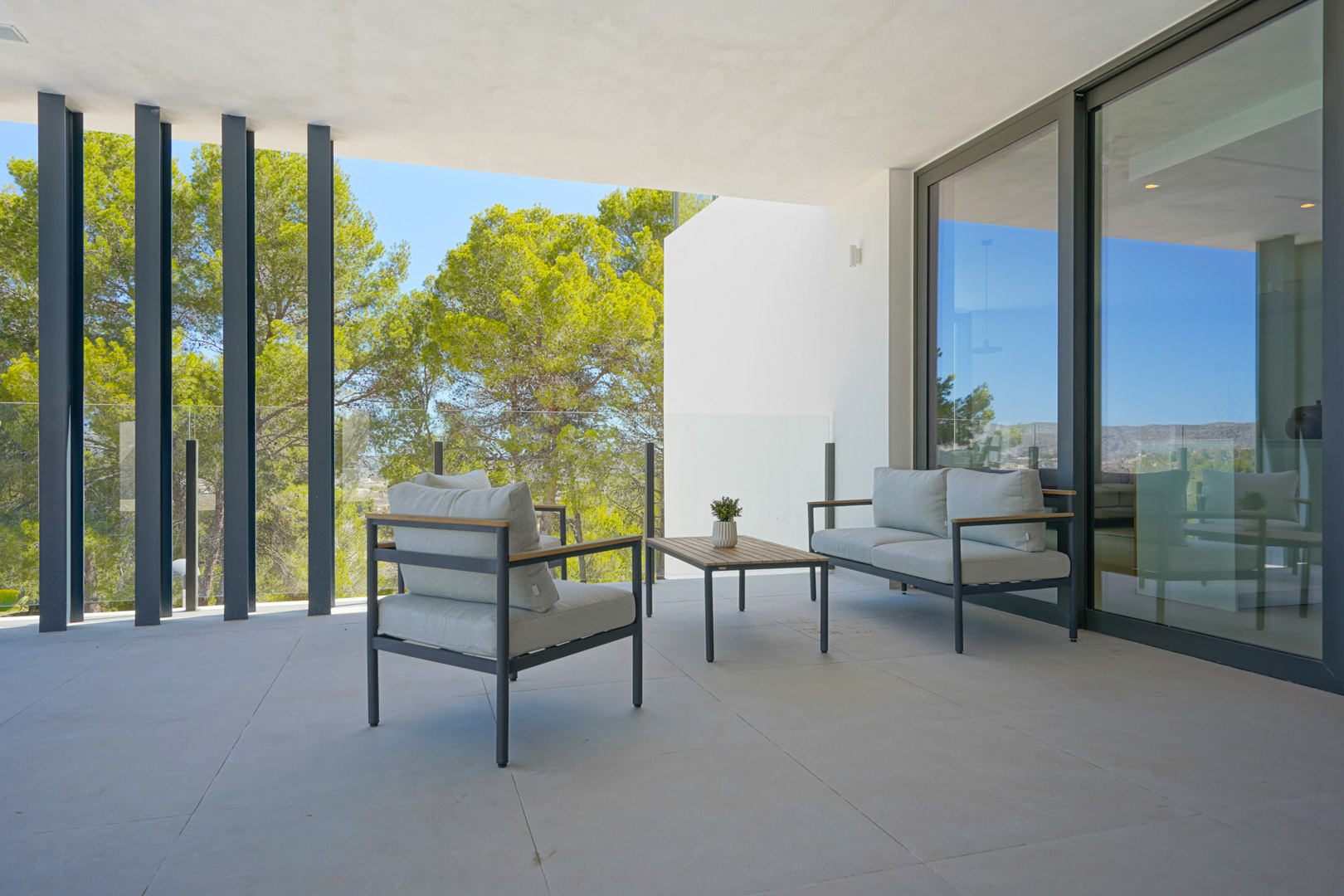 Covered terrace featuring modern lounge furniture, glass balustrades, and views of the pine forest through sleek vertical architectural elements.