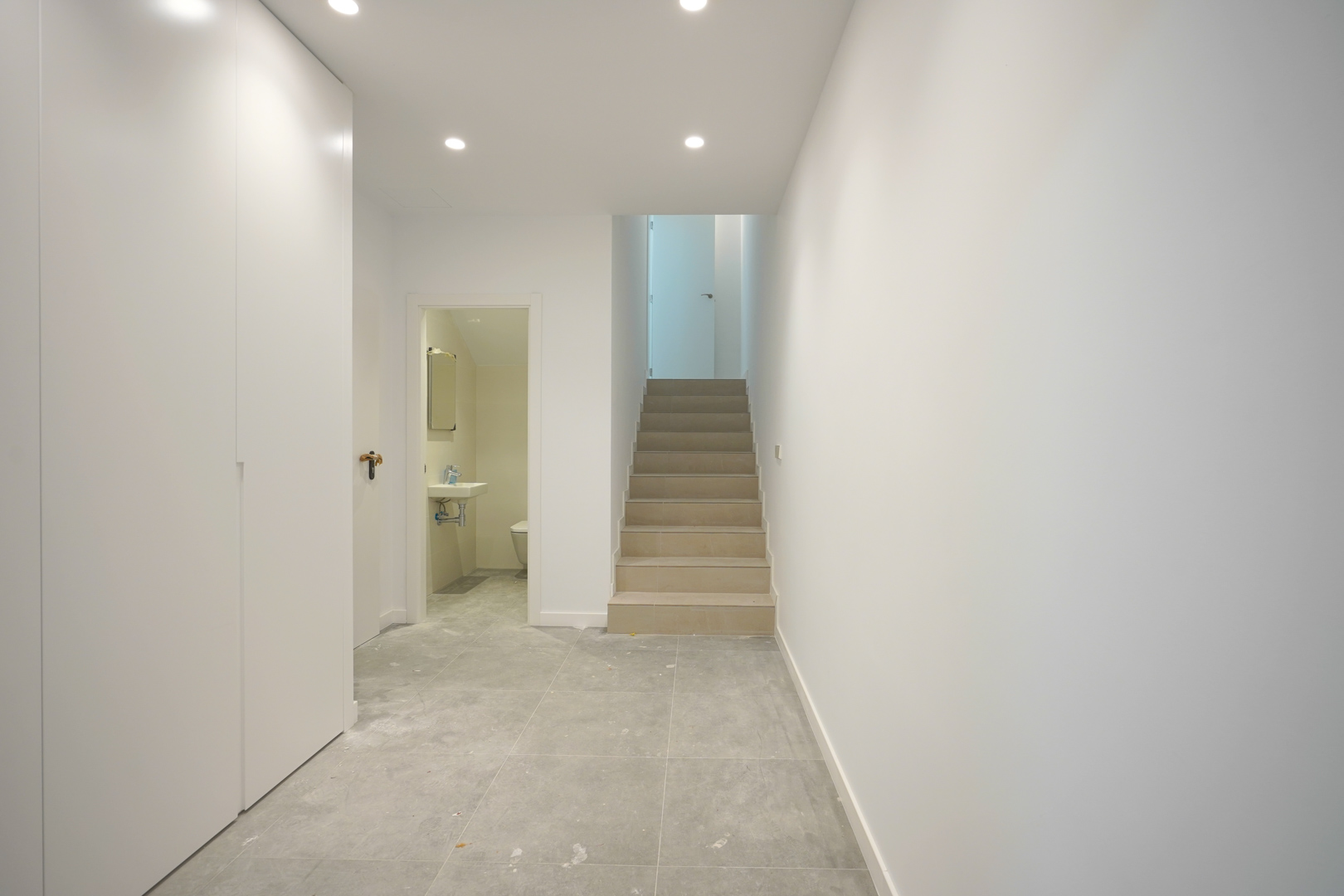 Distribution hallway featuring grey tiled flooring, white minimalist cabinetry, and a stone staircase. View includes a guest toilet and recessed LED ceiling lighting.