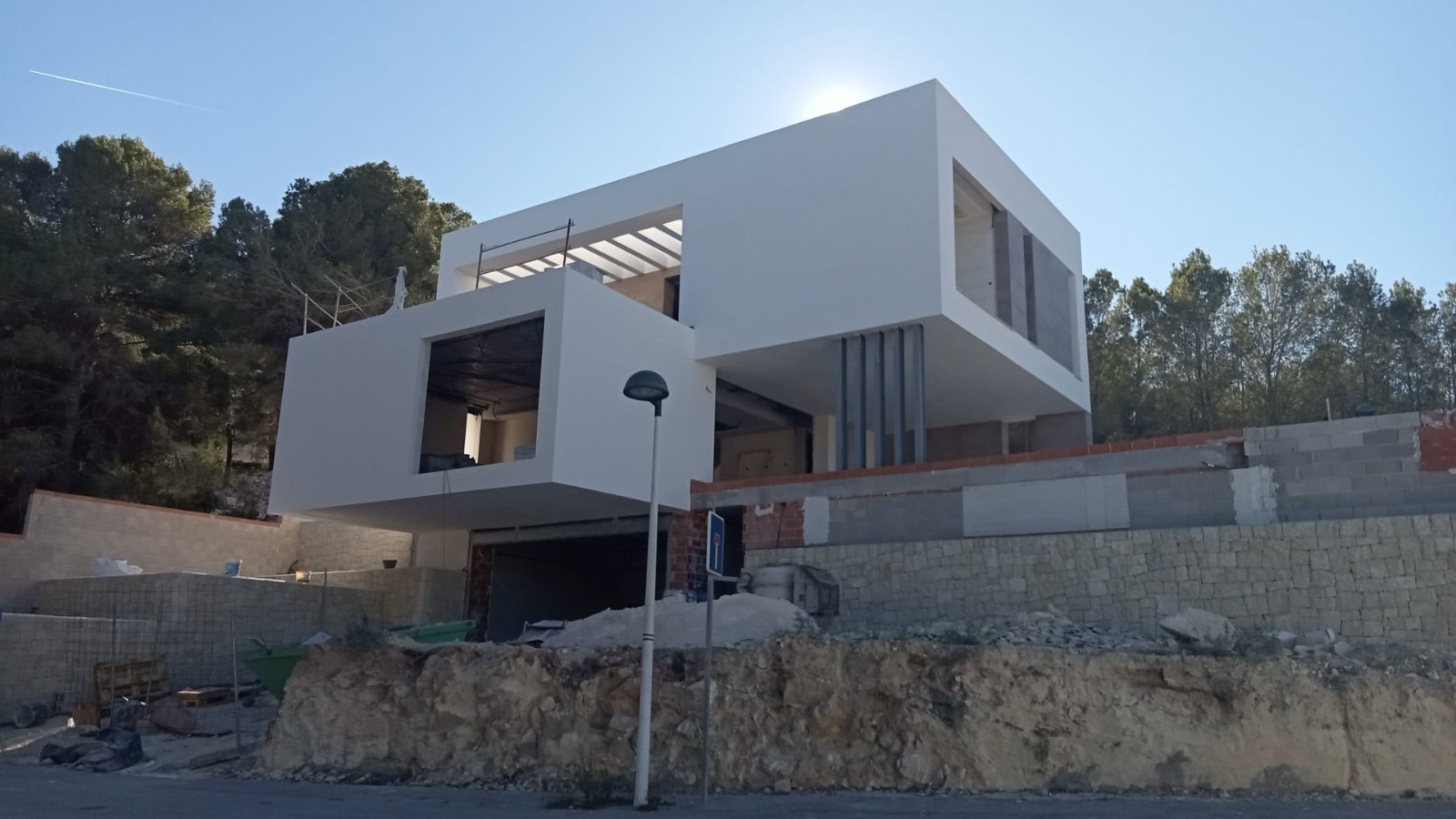 Exterior view of the villa under construction, showcasing minimalist cubist architecture with cantilevered volumes and a natural stone retaining wall.