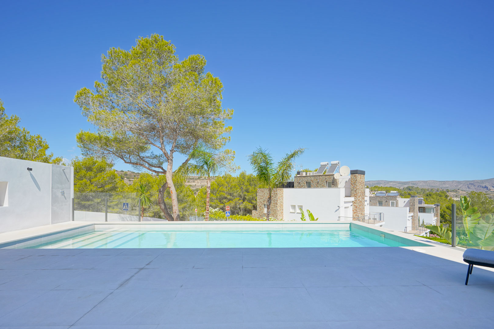 Infinity pool featuring white tiling and a large-format stone terrace, bordered by glass railings with views of Mediterranean pines and distant hills.