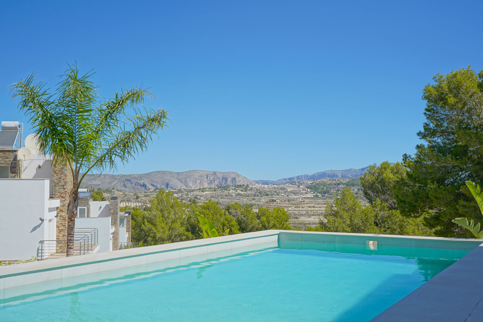 Infinity pool with white stone coping offering panoramic views of the mountains and valley, framed by Mediterranean pines and palm trees.