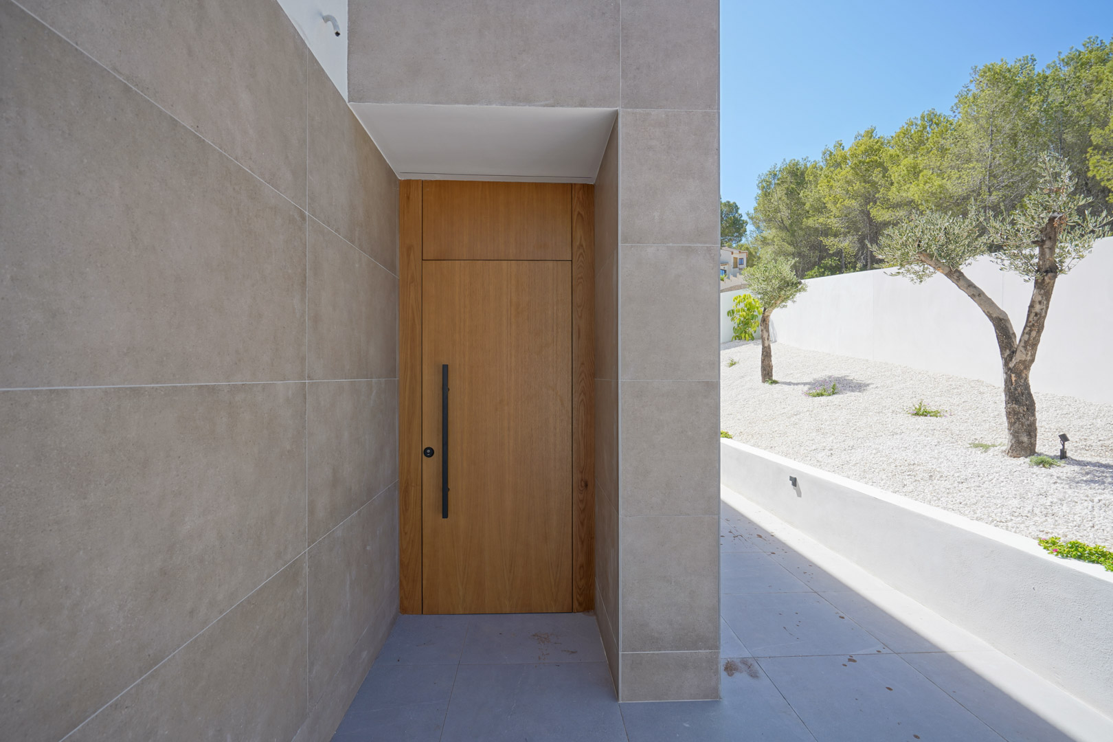 Main entrance featuring a solid wood door with a black handle, set against porcelain tile walls and a minimalist garden with olive trees.