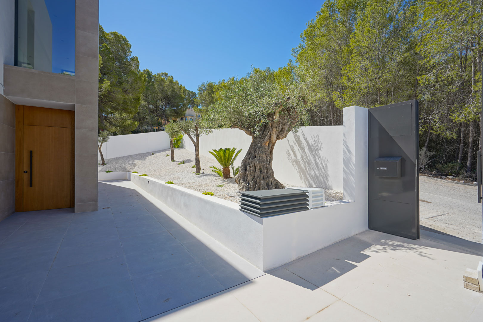 Main entrance featuring a wooden door, low-maintenance white gravel garden with olive trees, and a grey metal pedestrian gate.