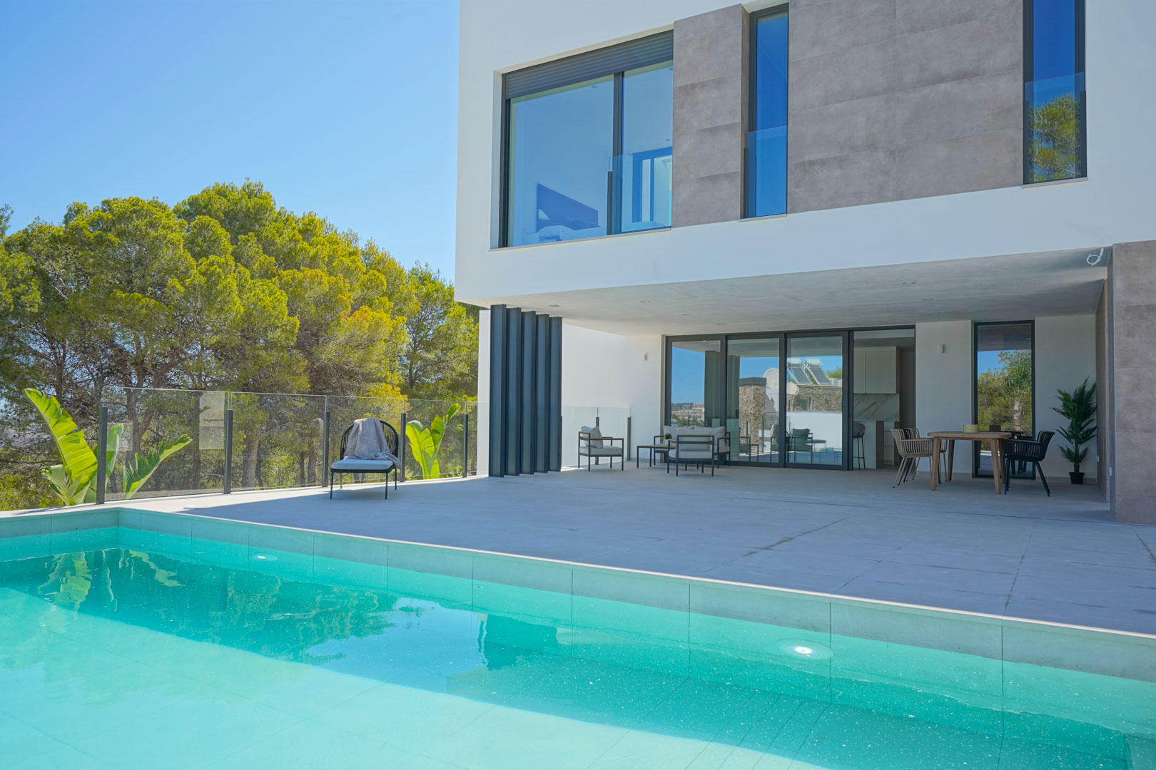 Main terrace by the swimming pool featuring outdoor lounge and dining furniture, set against a modern white facade and lush pine forest backdrop.
