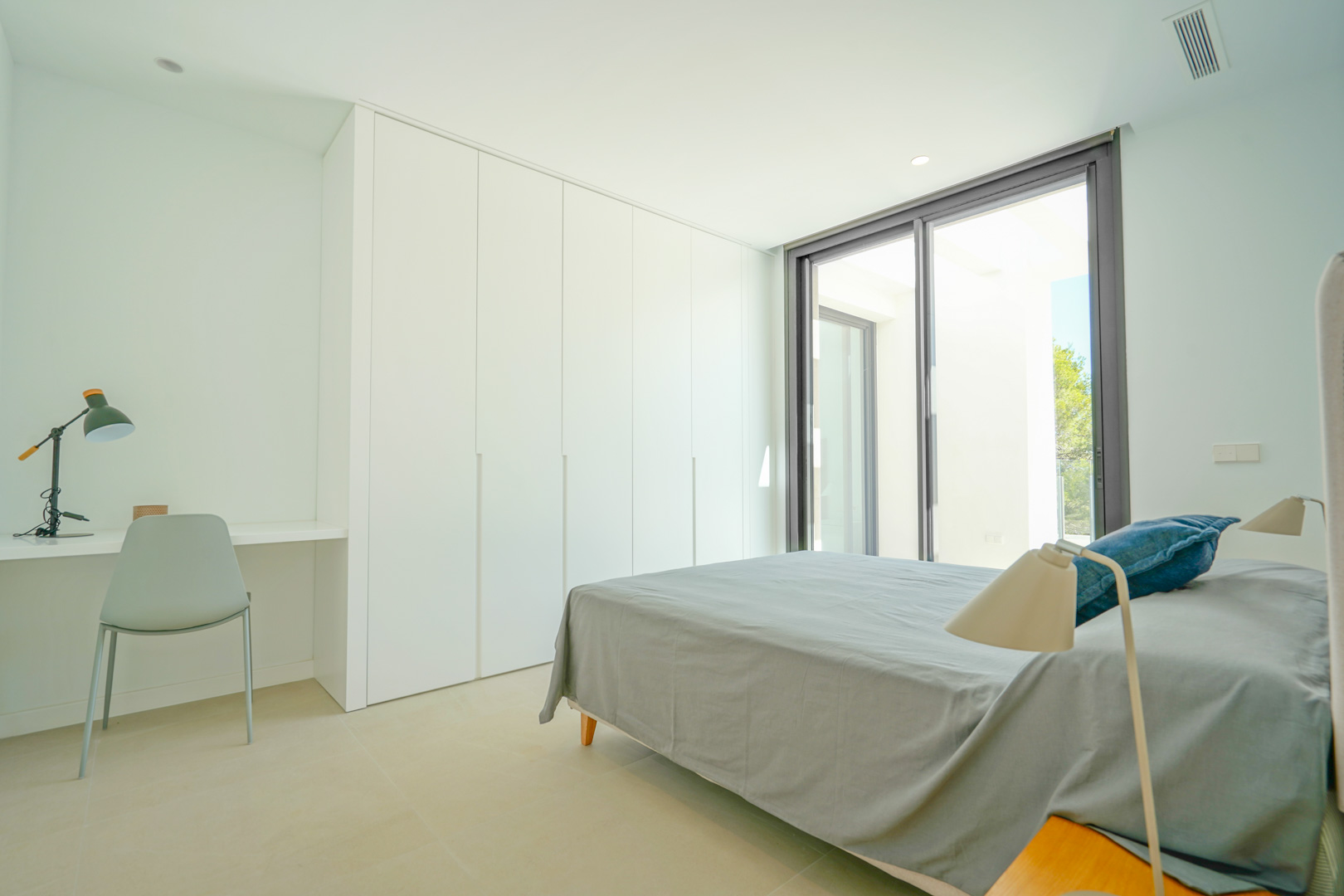 Minimalist bedroom featuring floor-to-ceiling white wardrobes, an integrated desk, and direct terrace access through large sliding glass doors.