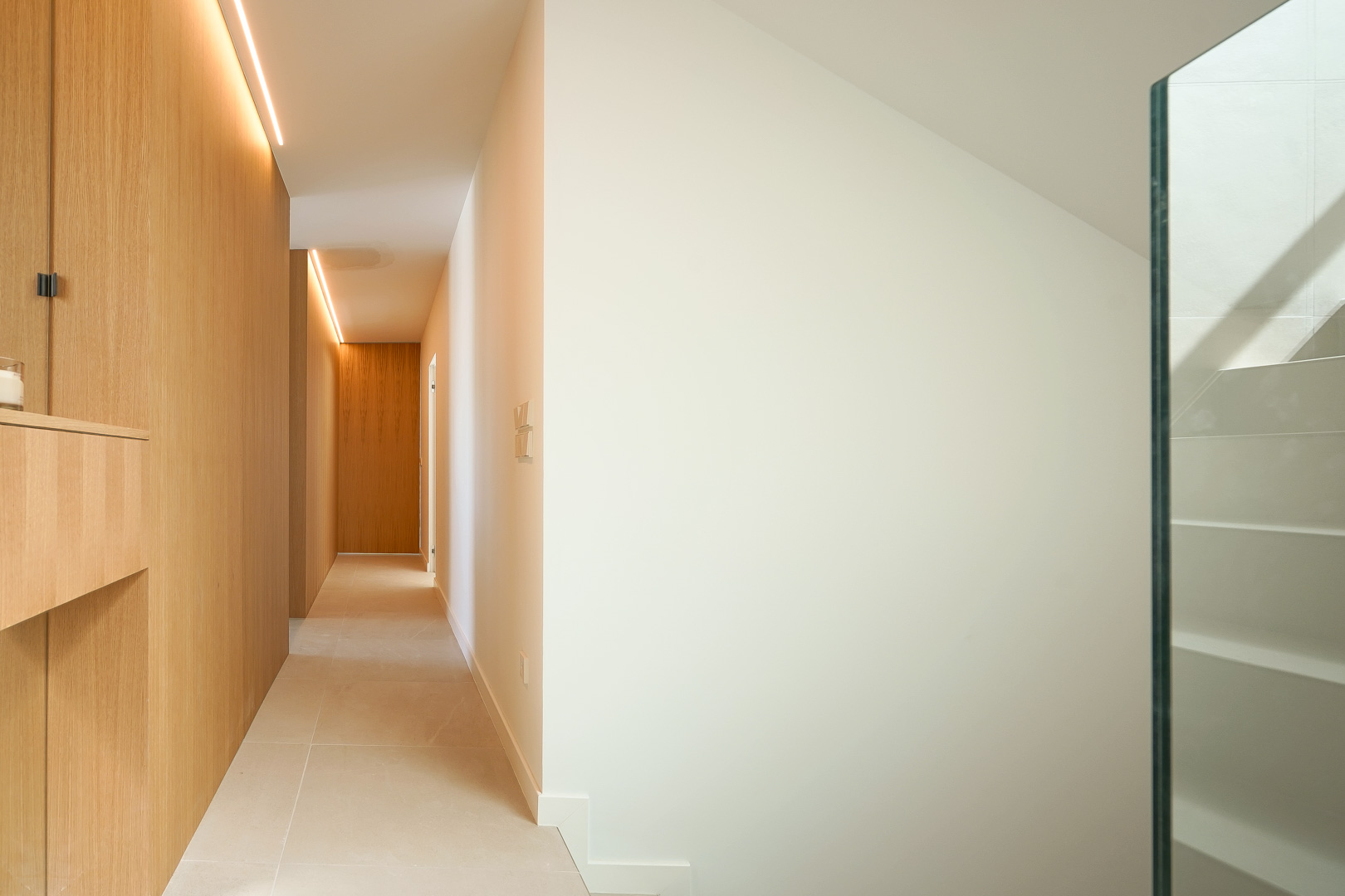 Minimalist hallway featuring oak wood paneling with integrated LED strip lighting, light porcelain flooring, and a staircase with a glass railing.