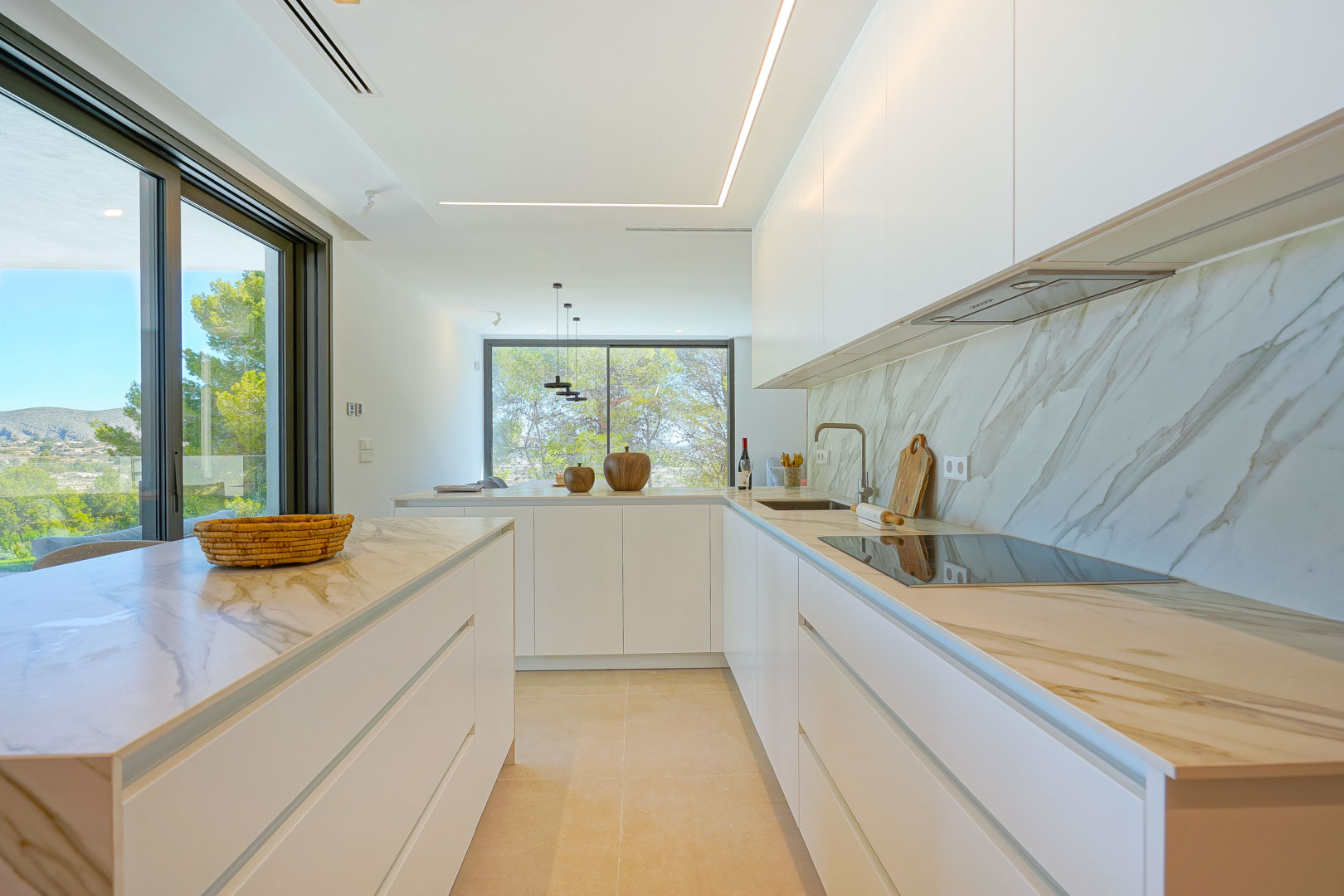 Minimalist open-plan kitchen featuring marble-effect countertops, handleless white cabinetry, and linear LED lighting. Large windows integrate views of the surrounding trees.
