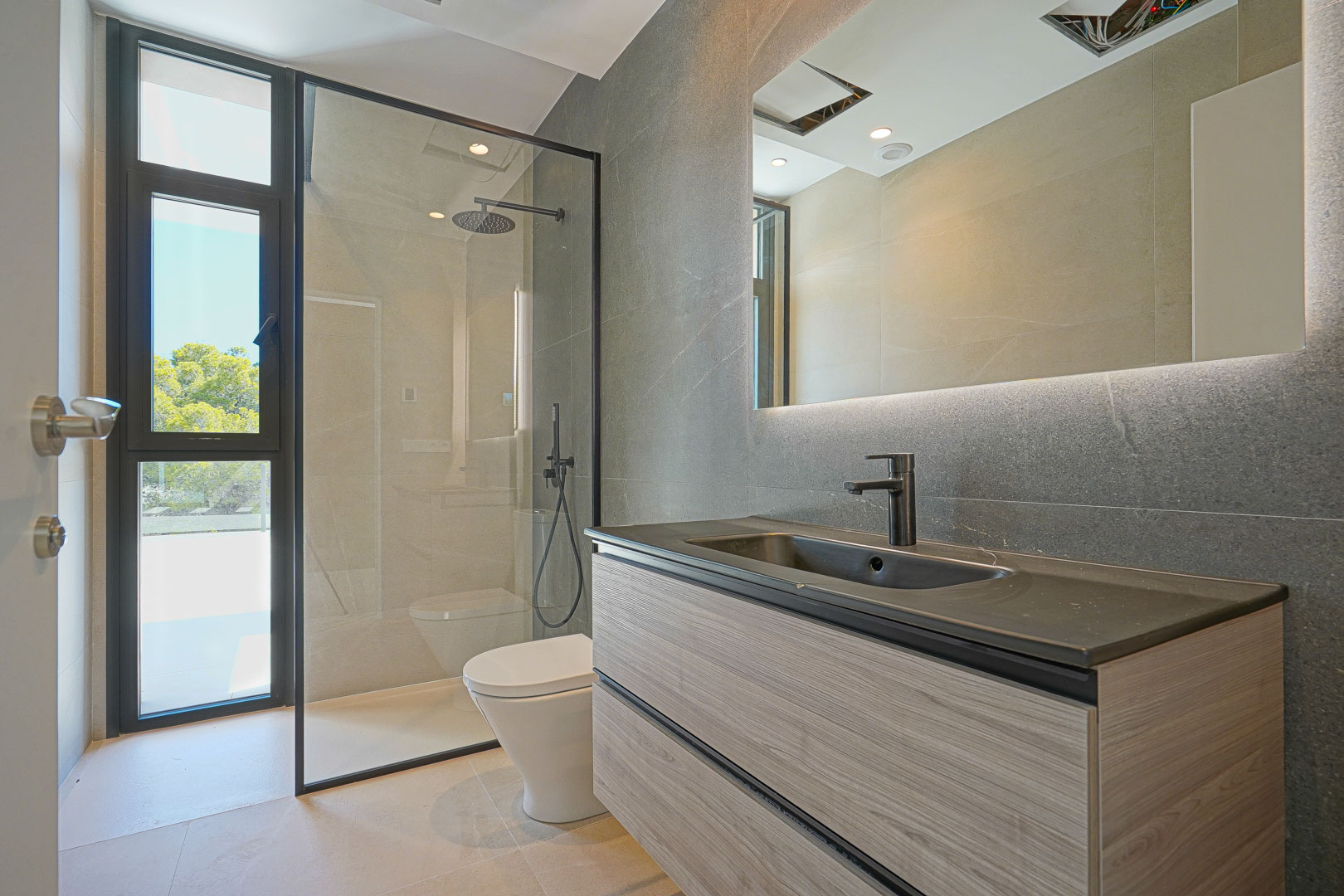 Modern bathroom featuring a floating wood-grain vanity, dark integrated sink, backlit mirror, and a walk-in shower with black-framed glass and natural light.