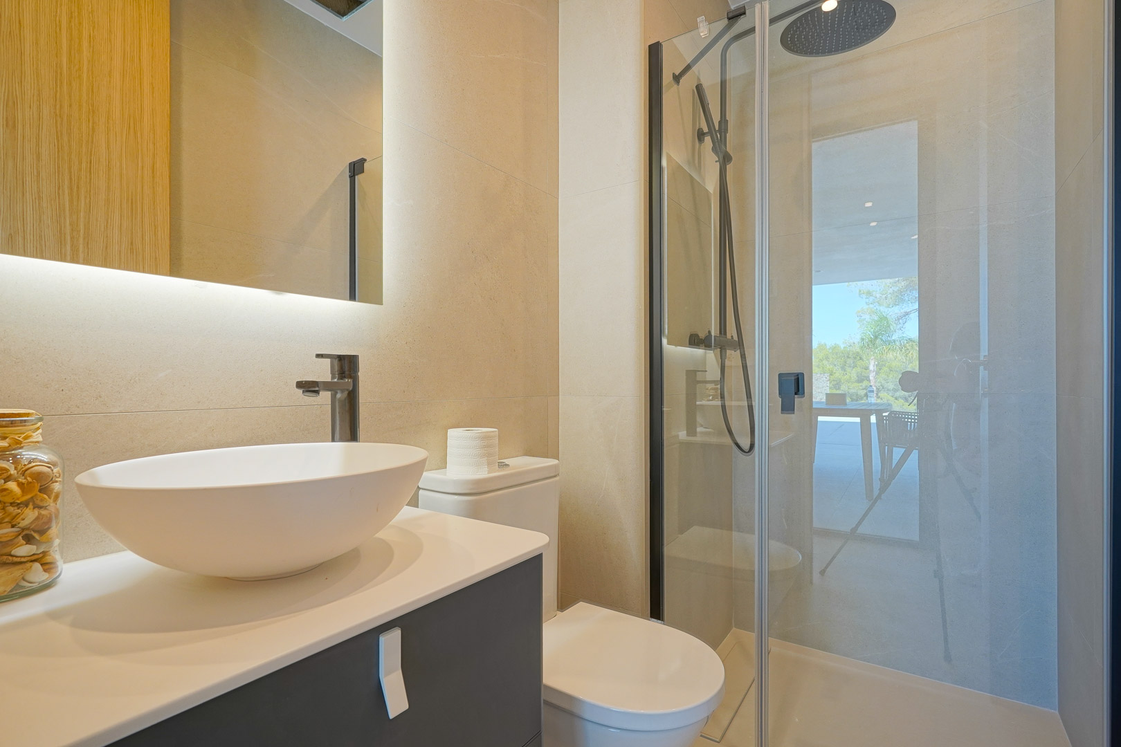 Modern bathroom featuring a vessel sink, backlit mirror, and walk-in shower with black hardware. Neutral beige tiling and natural light create a clean, contemporary space.