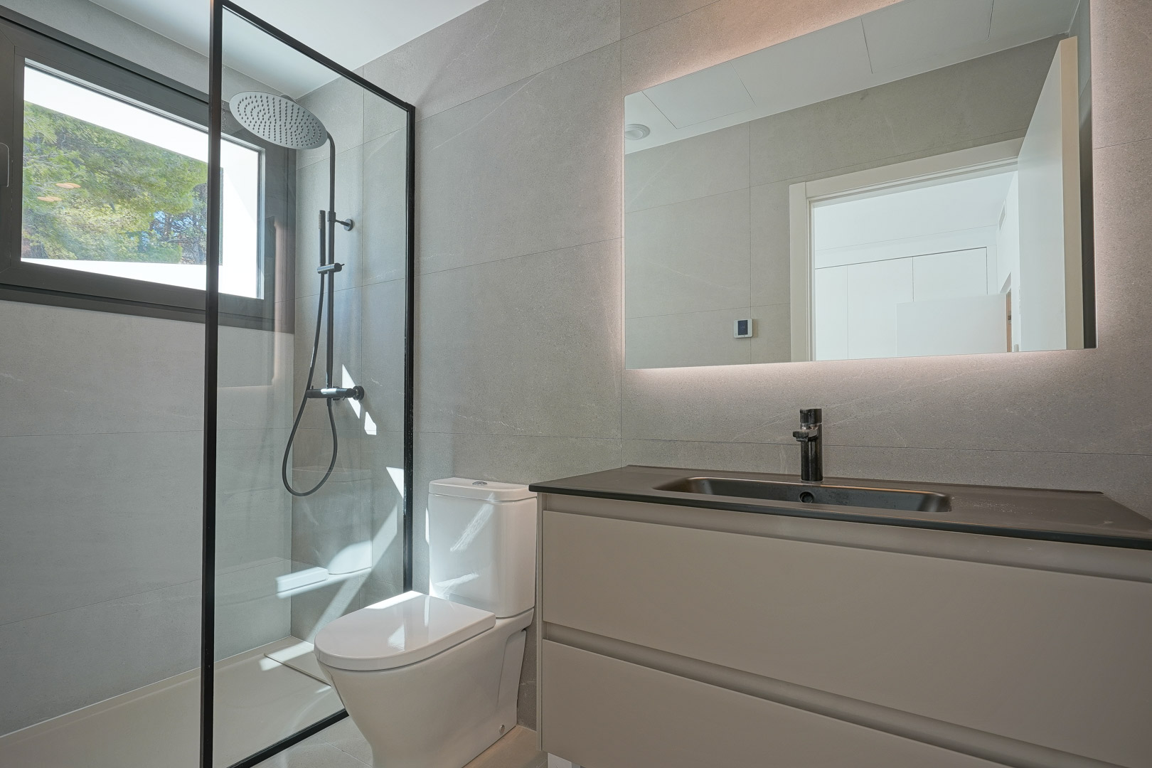 Modern bathroom featuring grey stone tiles, a walk-in shower with black framing, and a backlit mirror. It includes a sleek vanity with a dark countertop and integrated sink.