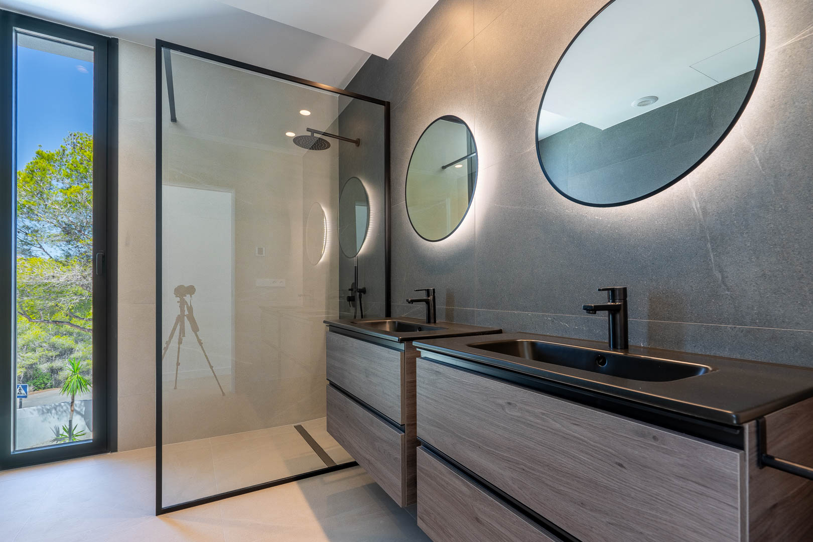 Modern bathroom with a double wooden vanity, backlit circular mirrors, and a walk-in shower featuring black hardware and large format tiles.