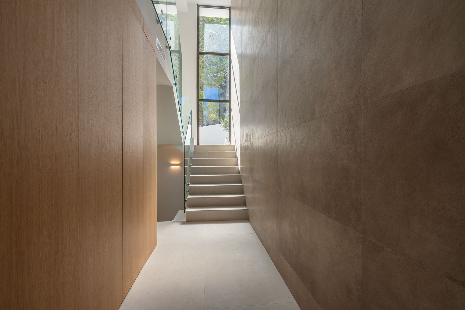 Modern hallway featuring wood paneling, large-format stone wall tiles, and a staircase with glass balustrades illuminated by a tall vertical window.