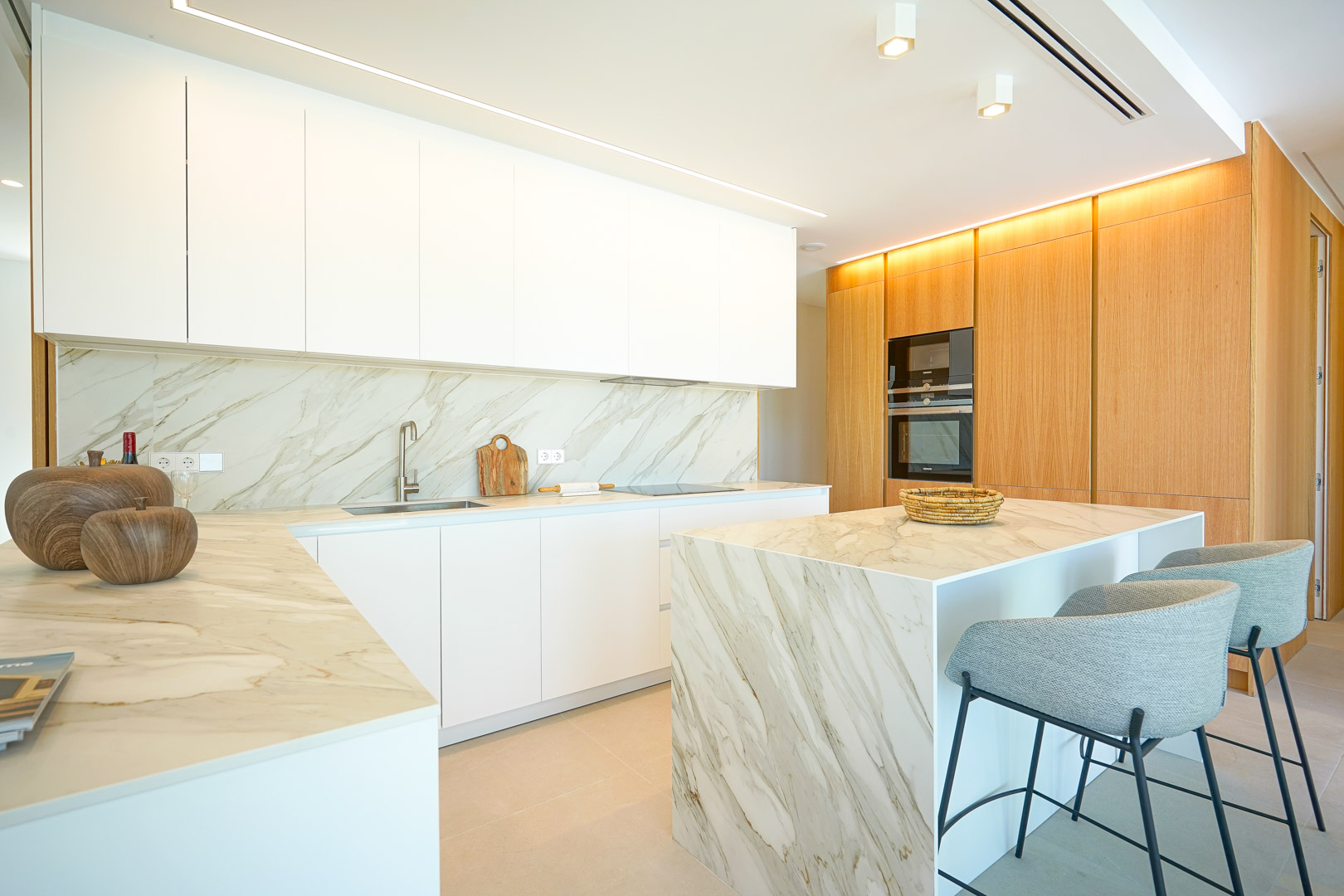 Modern kitchen featuring white handleless cabinetry, marble-effect countertops, and a matching island. A wooden wall integrates high-end appliances under recessed LED lighting.