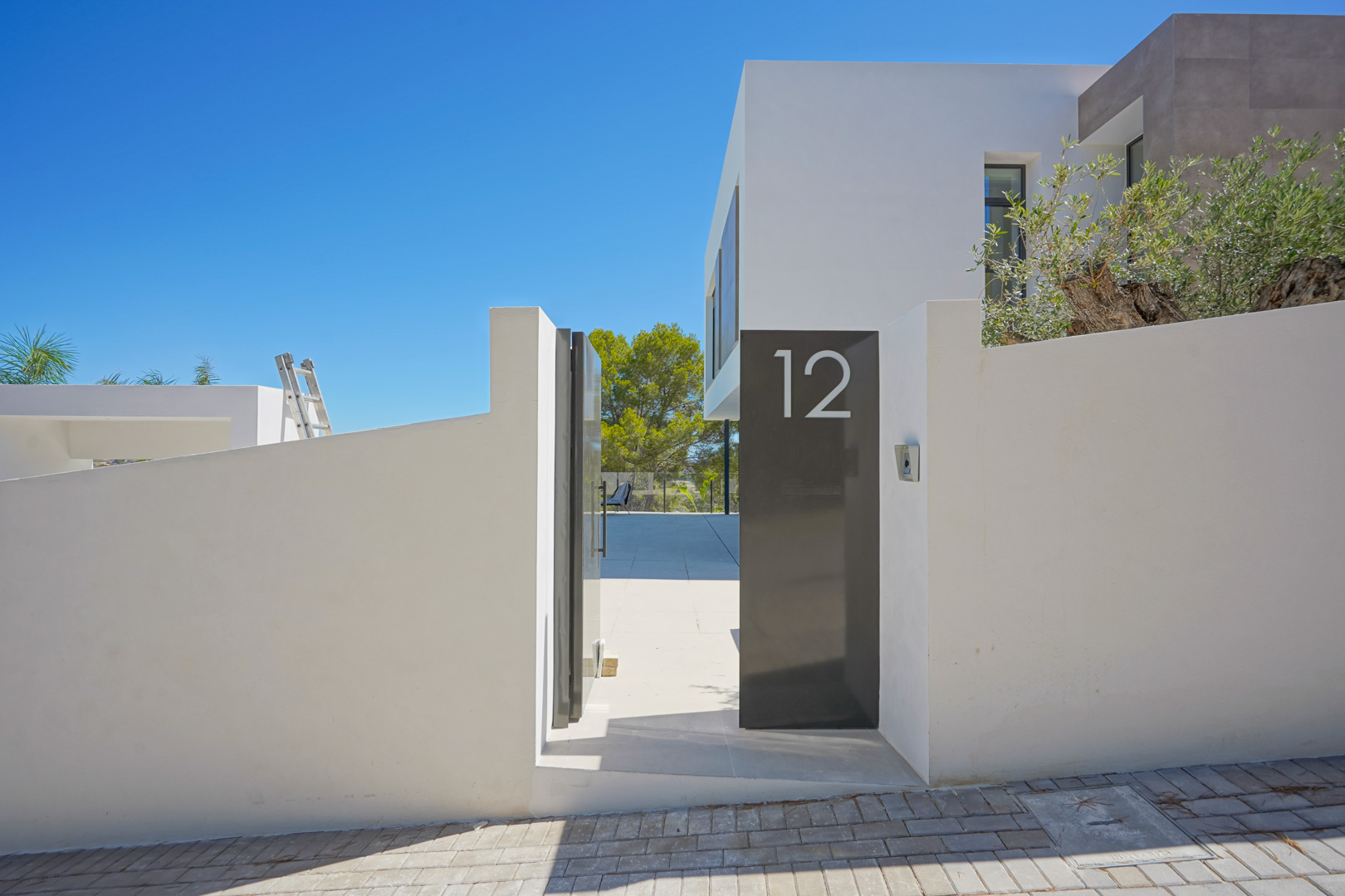 Modern main entrance featuring a minimalist white wall, dark grey metal pedestrian gate with house number 12, and a paved pathway leading into the property.