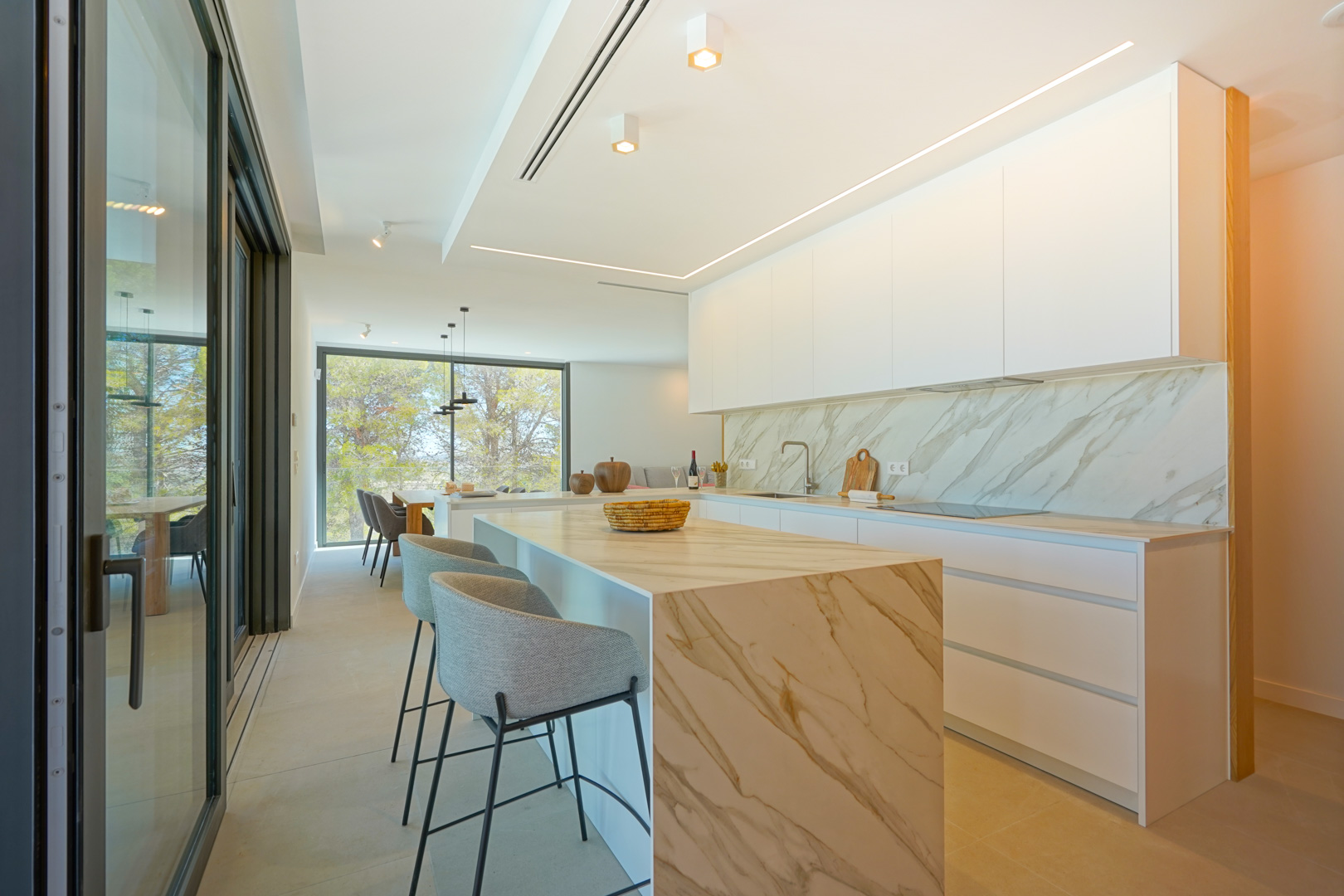 Modern open-plan kitchen with a marble-effect waterfall island and minimalist white cabinetry. Features integrated LED lighting and large sliding glass doors.