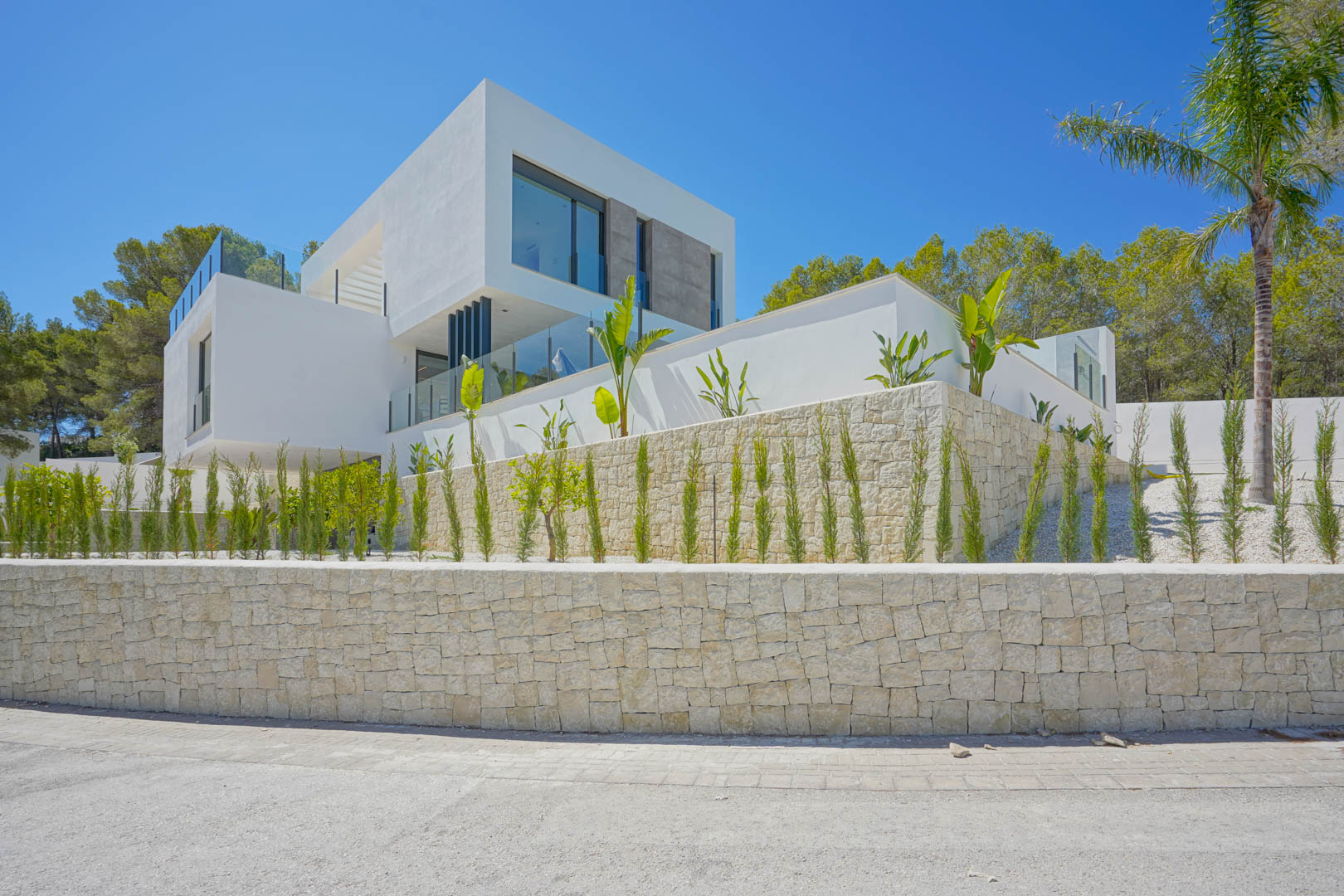 Modern villa facade featuring geometric white volumes, natural stone retaining walls, and landscaped gardens with young cypress trees and glass-railed terraces.