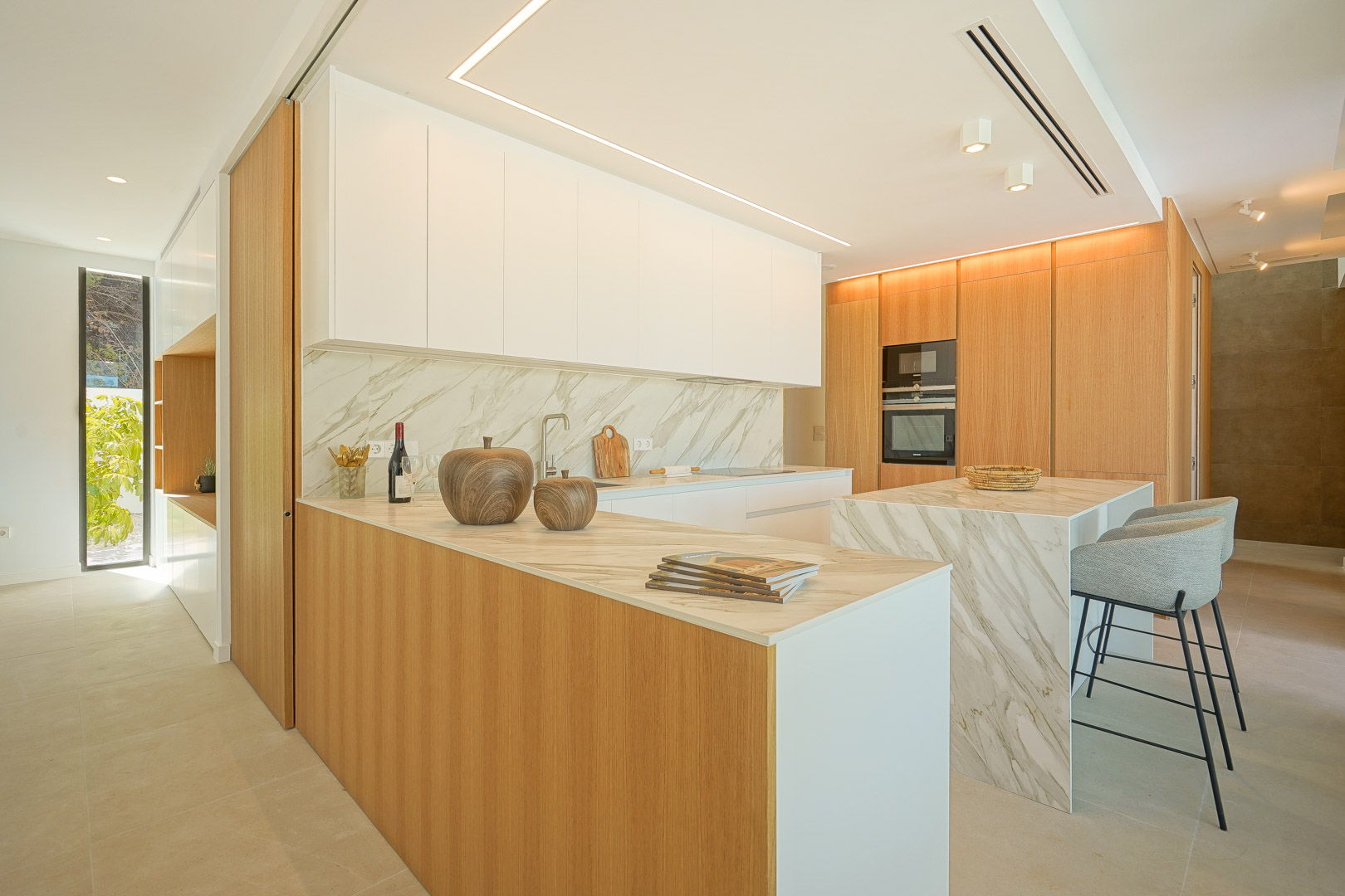 Open-plan kitchen featuring marble-effect countertops, oak wood cabinetry, and a central island with seating. Integrated appliances and linear LED ceiling lighting complete the space.