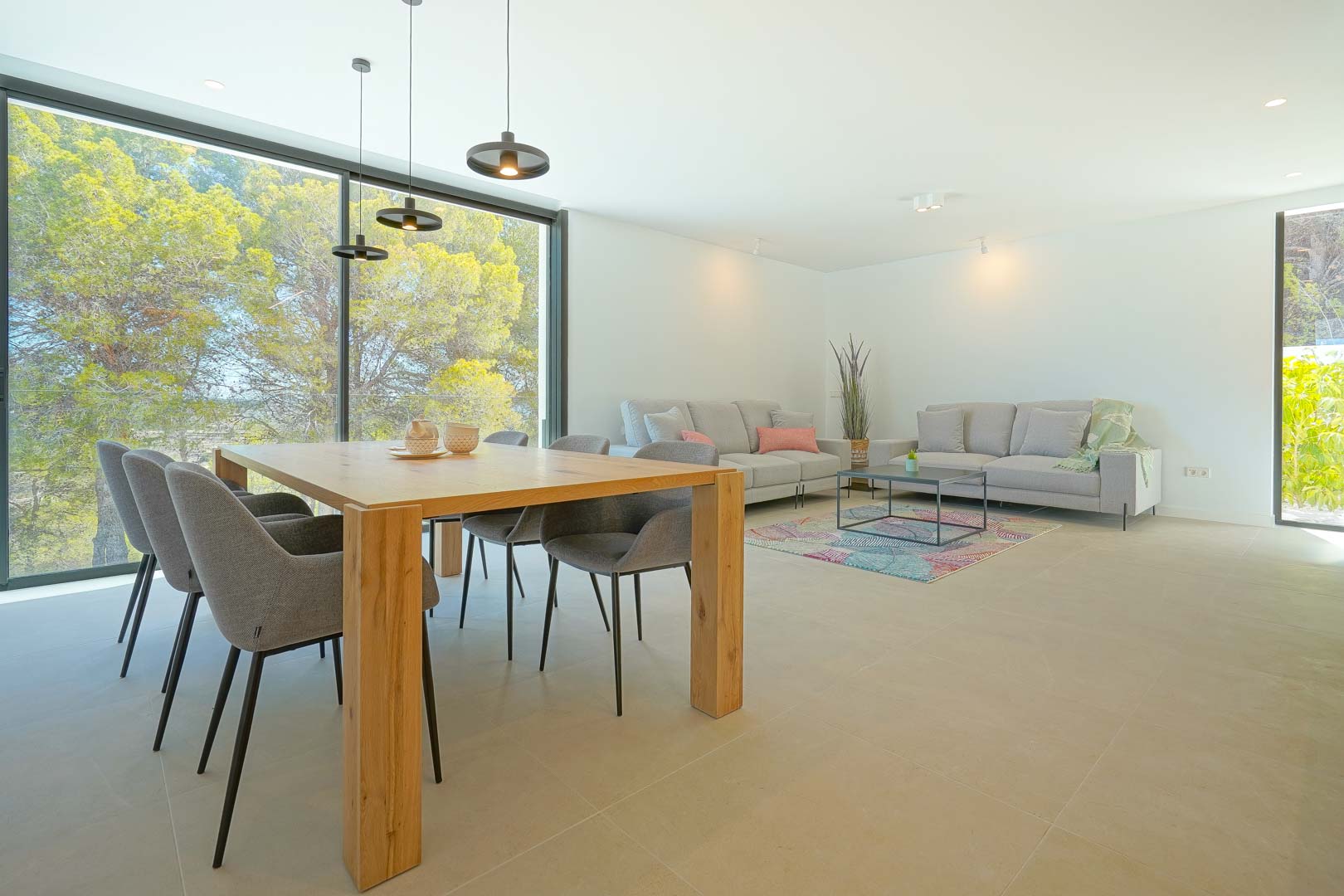 Open-plan living and dining area featuring a wooden table, grey upholstery, and floor-to-ceiling windows overlooking the surrounding pine forest.