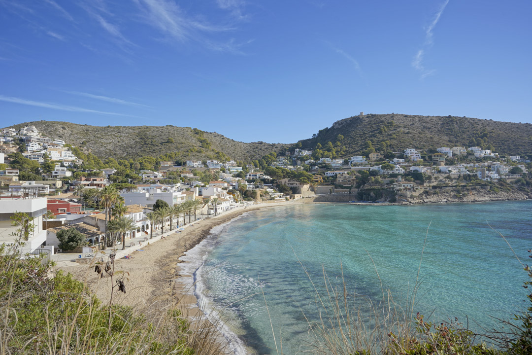 Panoramic view of El Portet beach in Moraira, featuring turquoise Mediterranean waters, a palm-lined promenade, and white villas nestled in the surrounding hills.