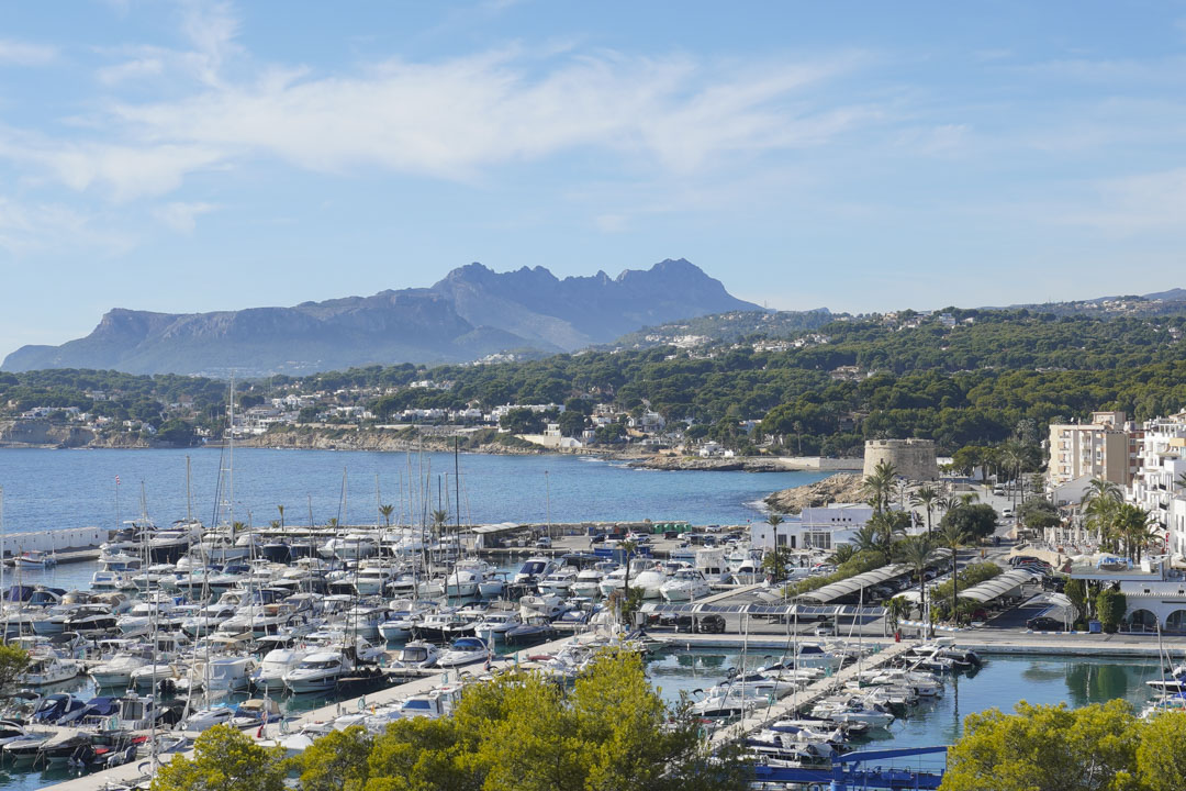 Panoramic views of Moraira's marina and historic watchtower, featuring the Bernia mountain range across the Mediterranean coastline.
