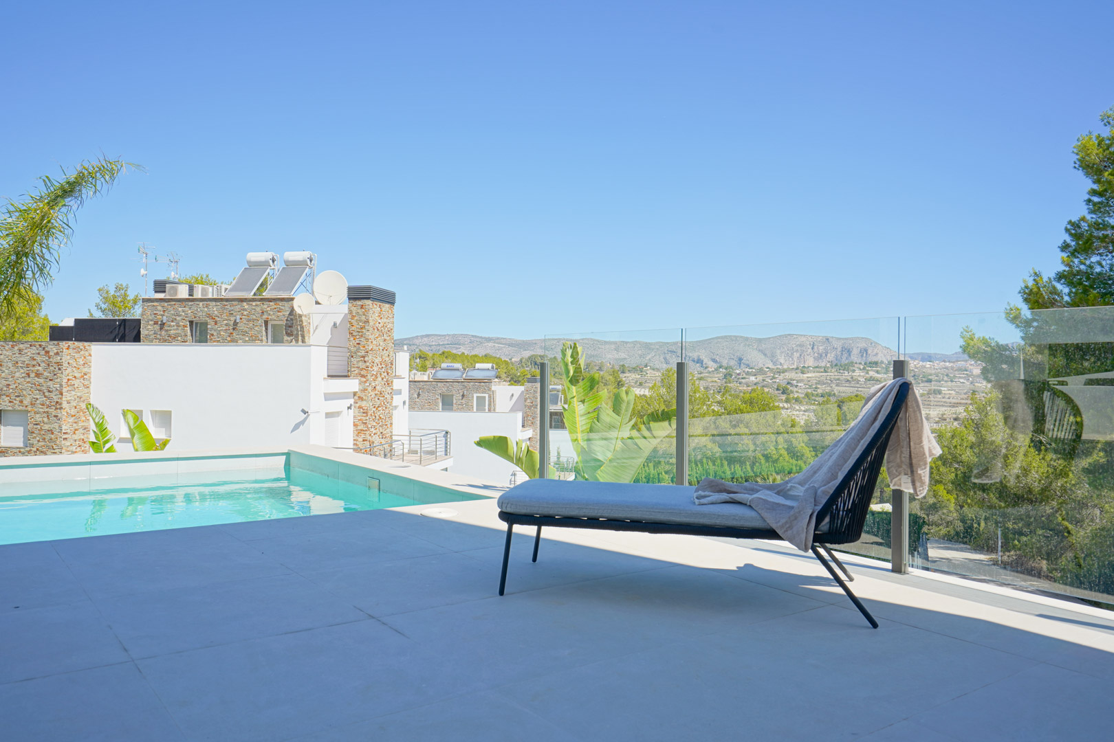 Poolside terrace featuring a glass balustrade, modern sun lounger, and panoramic views of the surrounding mountains and Mediterranean landscape.