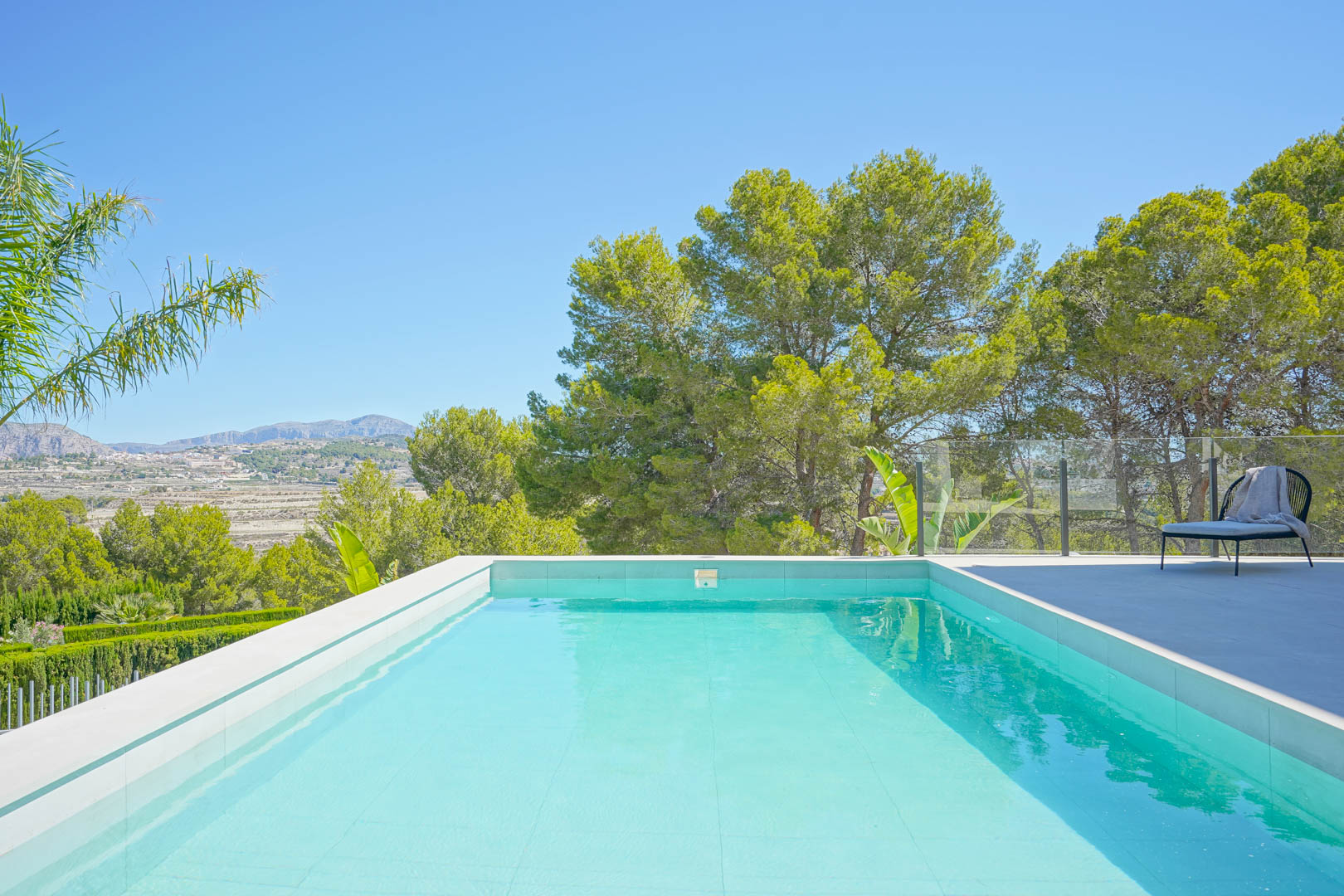 Rectangular swimming pool with light blue tiling, a glass balustrade, and a spacious terrace offering views of the Mediterranean pine forest and mountains.