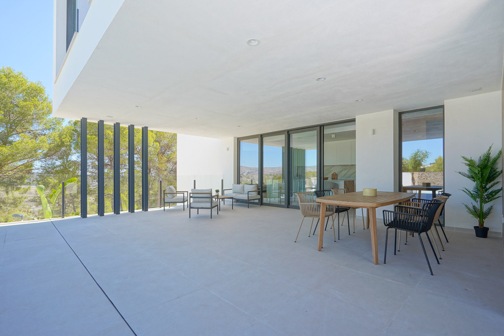 Spacious covered terrace featuring a wooden dining set, lounge furniture, and glass railings with views of the surrounding pine forest and mountains.