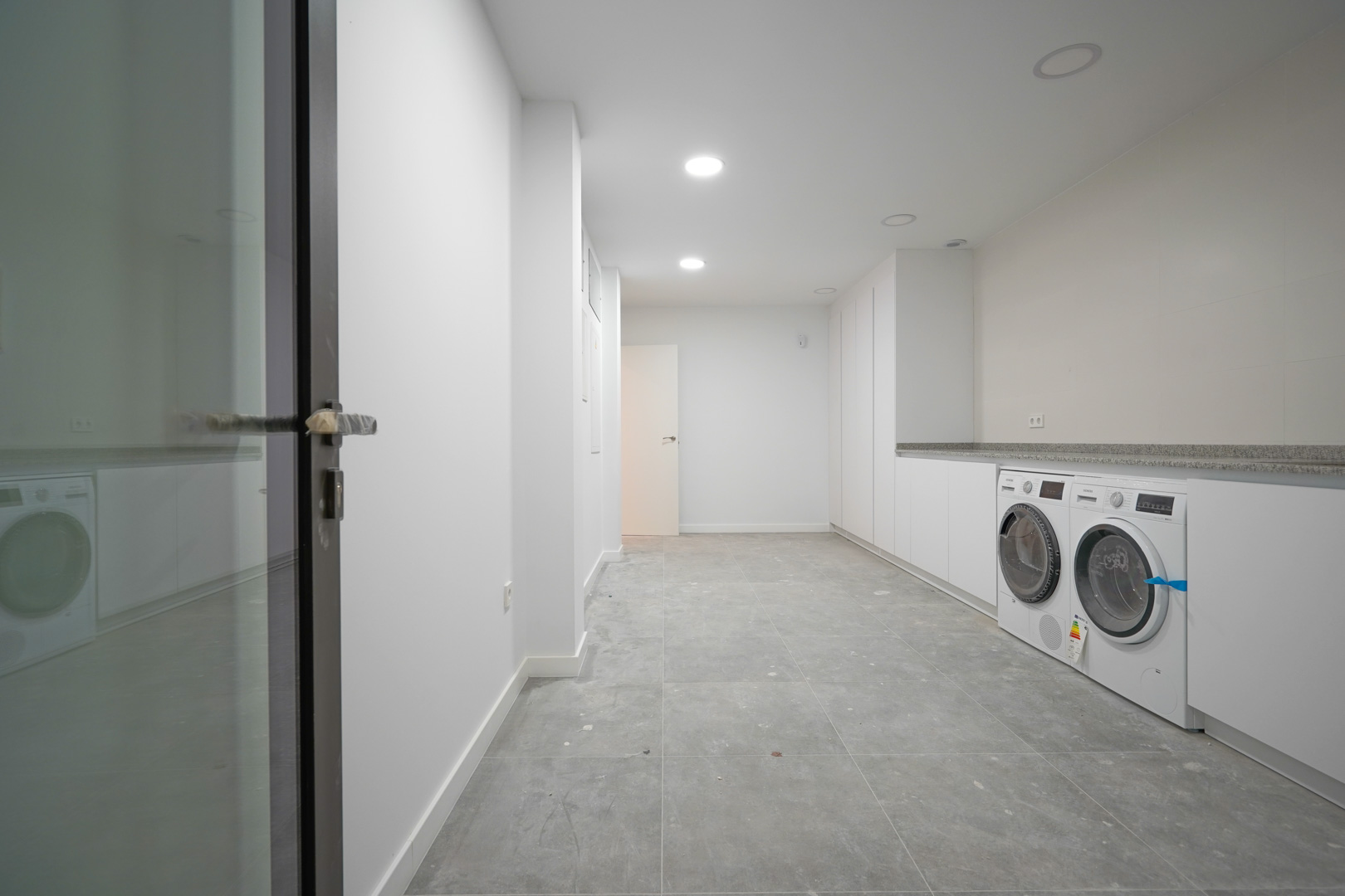 Spacious ground floor laundry room featuring white cabinetry, a stone countertop, integrated Siemens appliances, and grey tiled flooring.