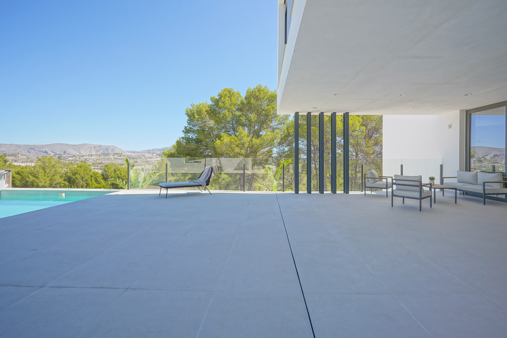 Spacious terrace featuring large-format tiling, a covered lounge area with minimalist furniture, and glass railings overlooking the pine forest.