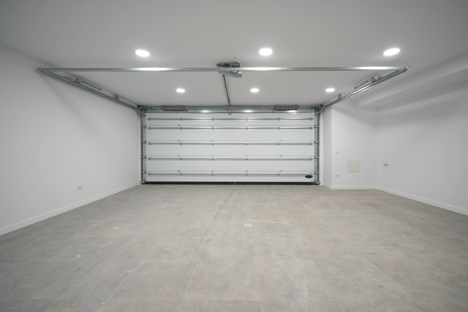 Spacious two-car garage featuring an automatic sectional door, grey tiled flooring, and recessed LED ceiling lighting.