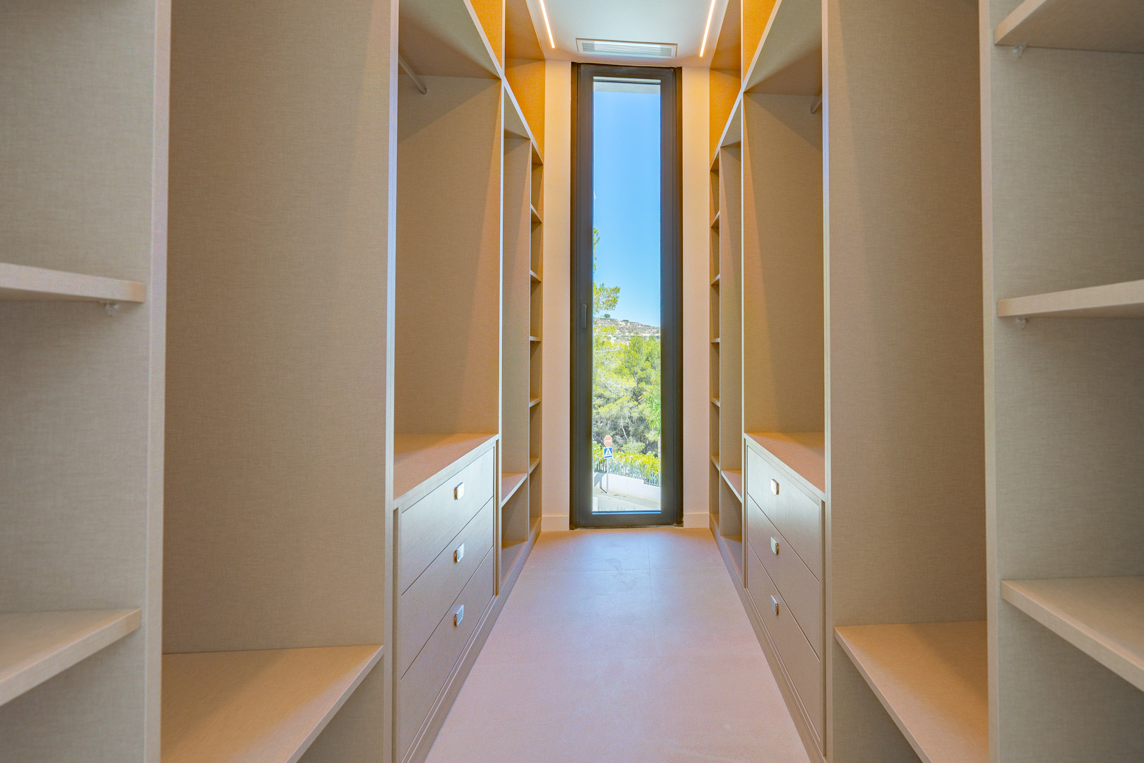 Spacious walk-in closet featuring minimalist shelving, integrated drawers, and a tall vertical window providing natural light and views of the greenery.