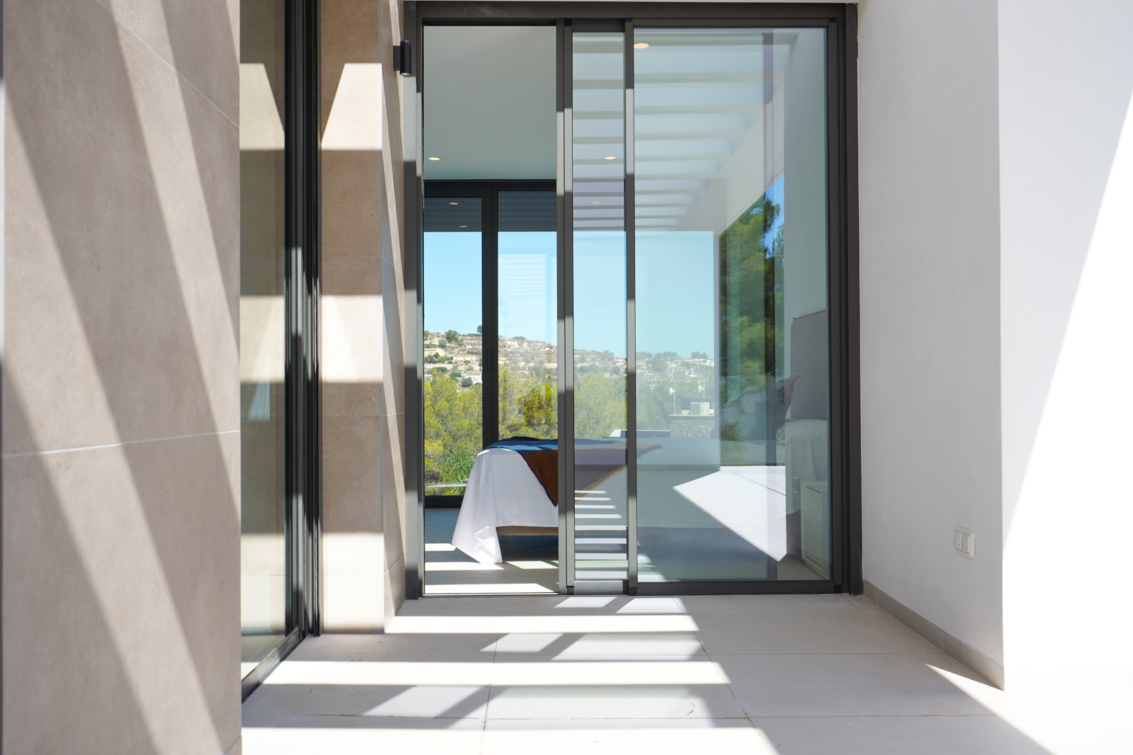Terrace view featuring stone cladding and sliding glass doors leading to a bright bedroom. Linear shadows from the pergola fall on the tiled floor.