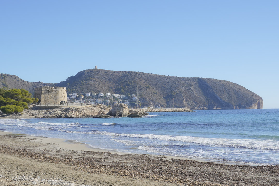 View of Moraira beach featuring the historic stone watchtower and Cap d'Or headland against a clear blue Mediterranean sky.
