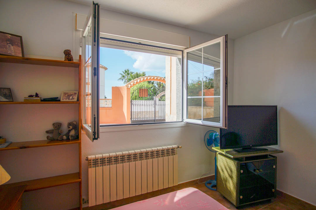 Bright bedroom featuring solid wood shelving, a radiator, and a window offering views of the gated entrance and Mediterranean greenery.