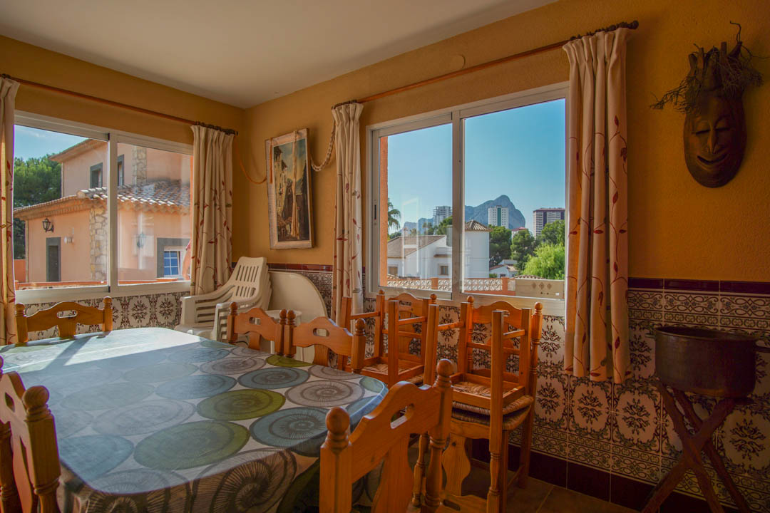 Bright dining area featuring solid wood furniture, traditional decorative wall tiles, and large windows offering views of the Peñón de Ifach.