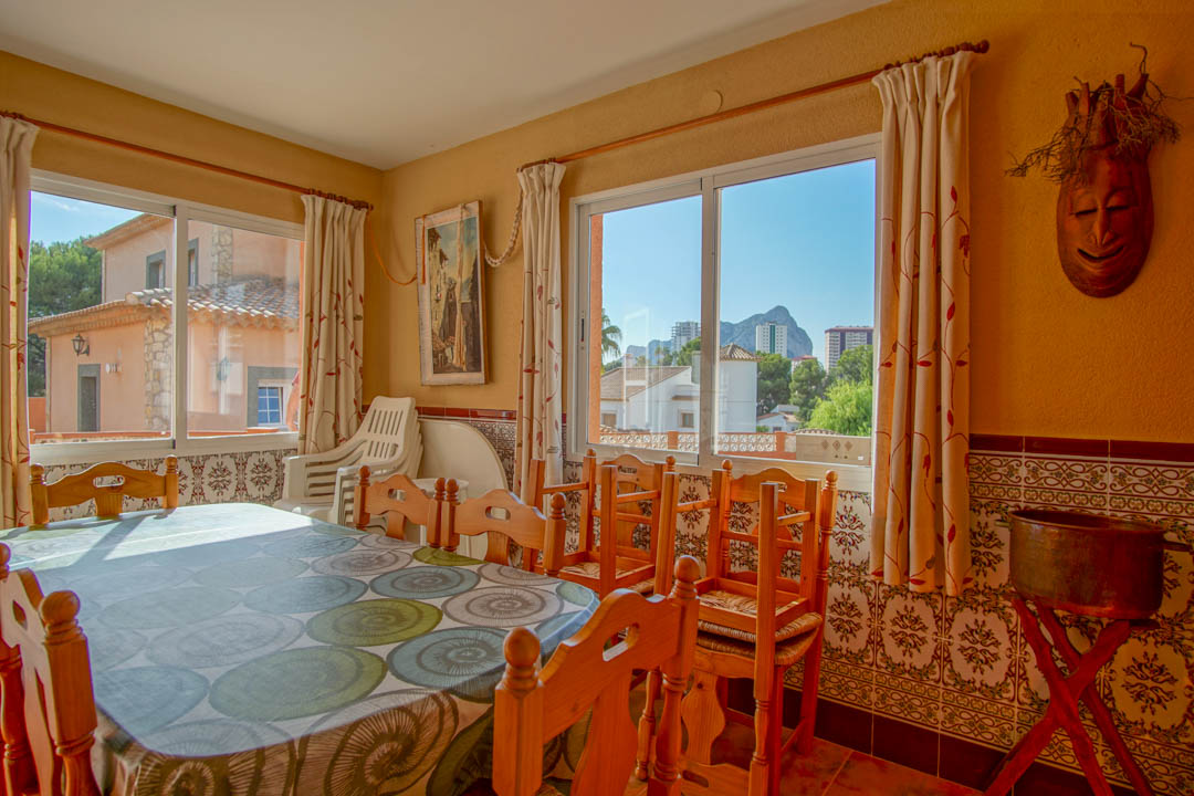 Bright dining area featuring solid wood furniture, traditional patterned wall tiles, and large windows with views of the Peñón de Ifach and the local skyline.