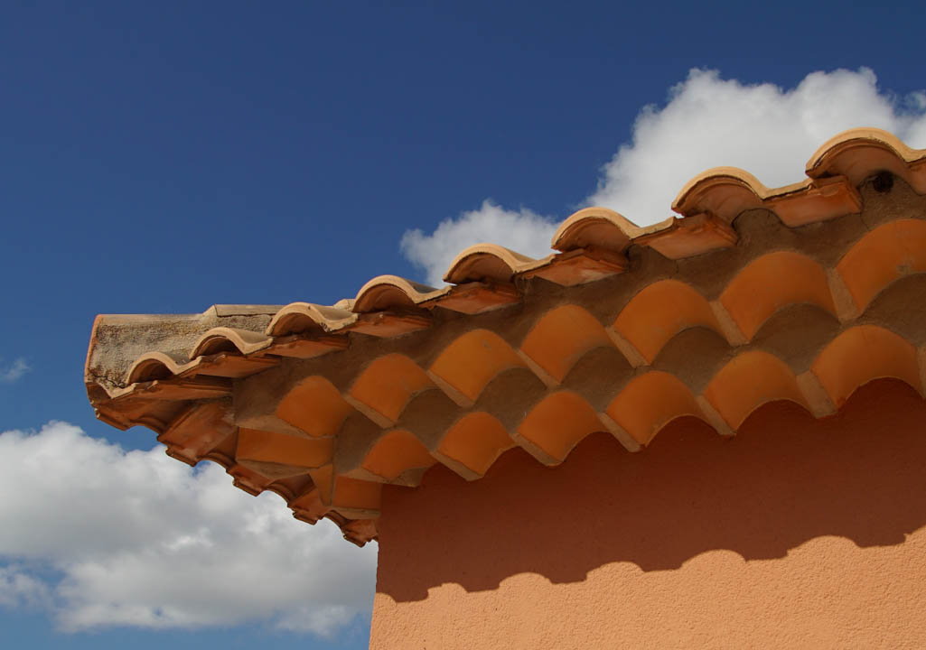 Close-up of traditional terracotta roof tiles featuring a decorative triple-layered cornice against a warm-toned exterior wall and blue sky.